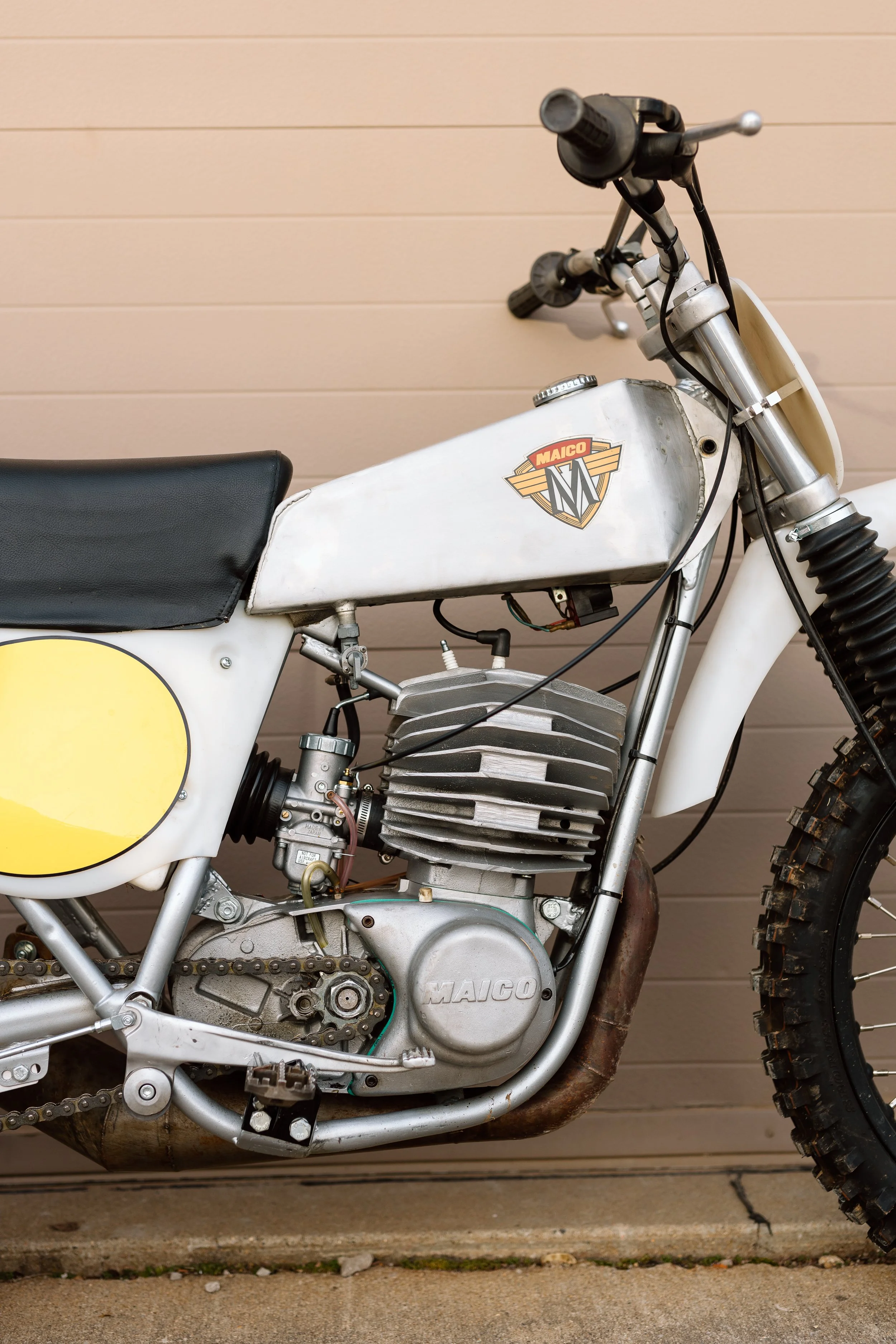 A vintage Maico dirt bike with a white frame, black seat, and visible engine, parked against a beige wall.
