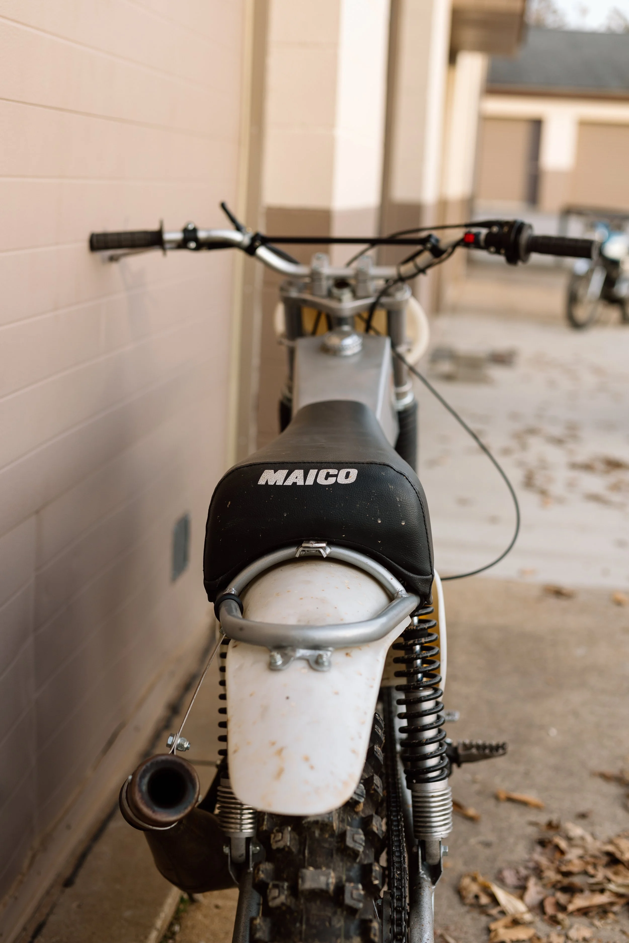 Rear view of a dirt bike with a black seat labeled MAICO, white fender, and worn tires, parked near a wall.