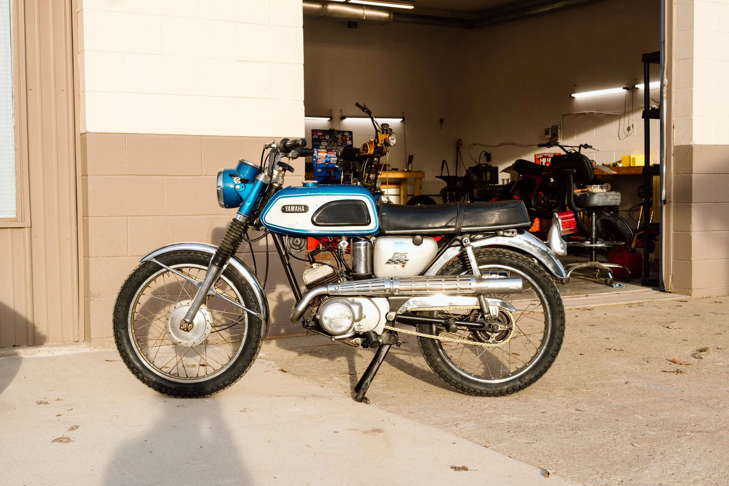 A vintage Yamaha motorcycle parked outside a garage with tools and equipment inside.