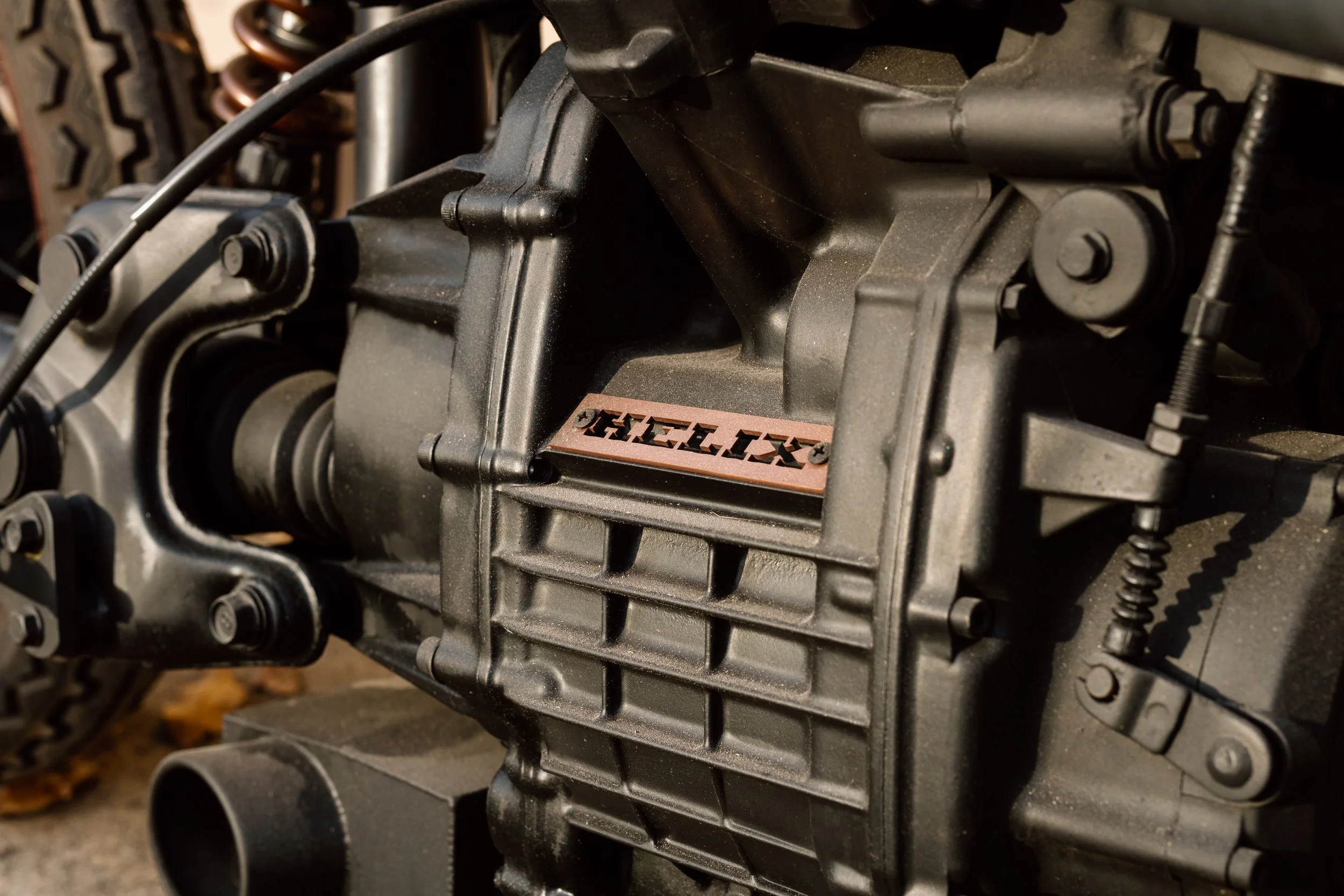 Close-up of a car engine, focusing on the black metal engine block with some visible bolts, a connected hose, and a small metallic component with the inscription 'OHEC.TX' on a copper-colored plate.