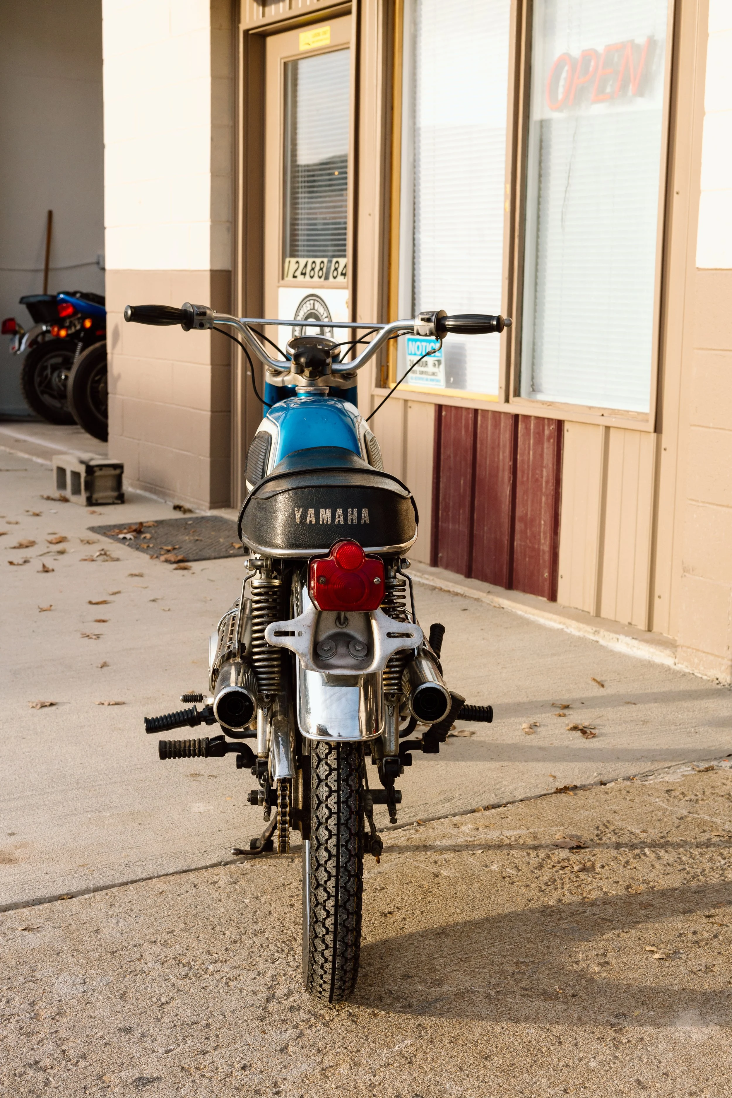 Rear view of a vintage Yamaha motorcycle parked outside a building with a glass door and window, with an open sign visible.