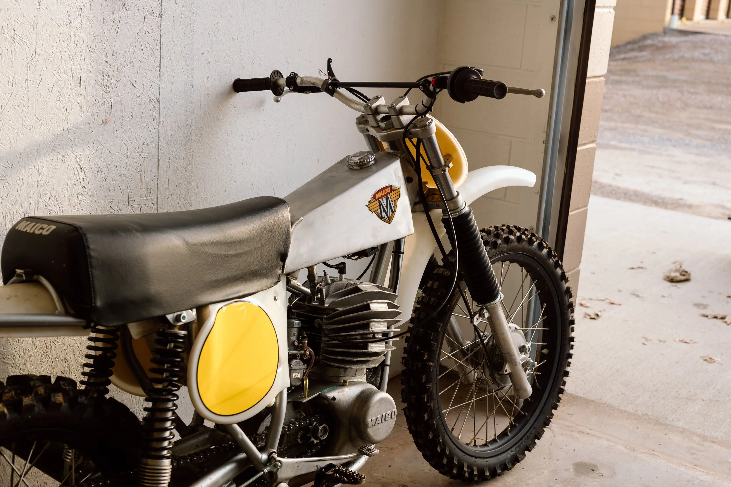 Vintage Maico dirt bike with black seat, yellow and white body, and knobby tires, parked inside a garage near an open door.