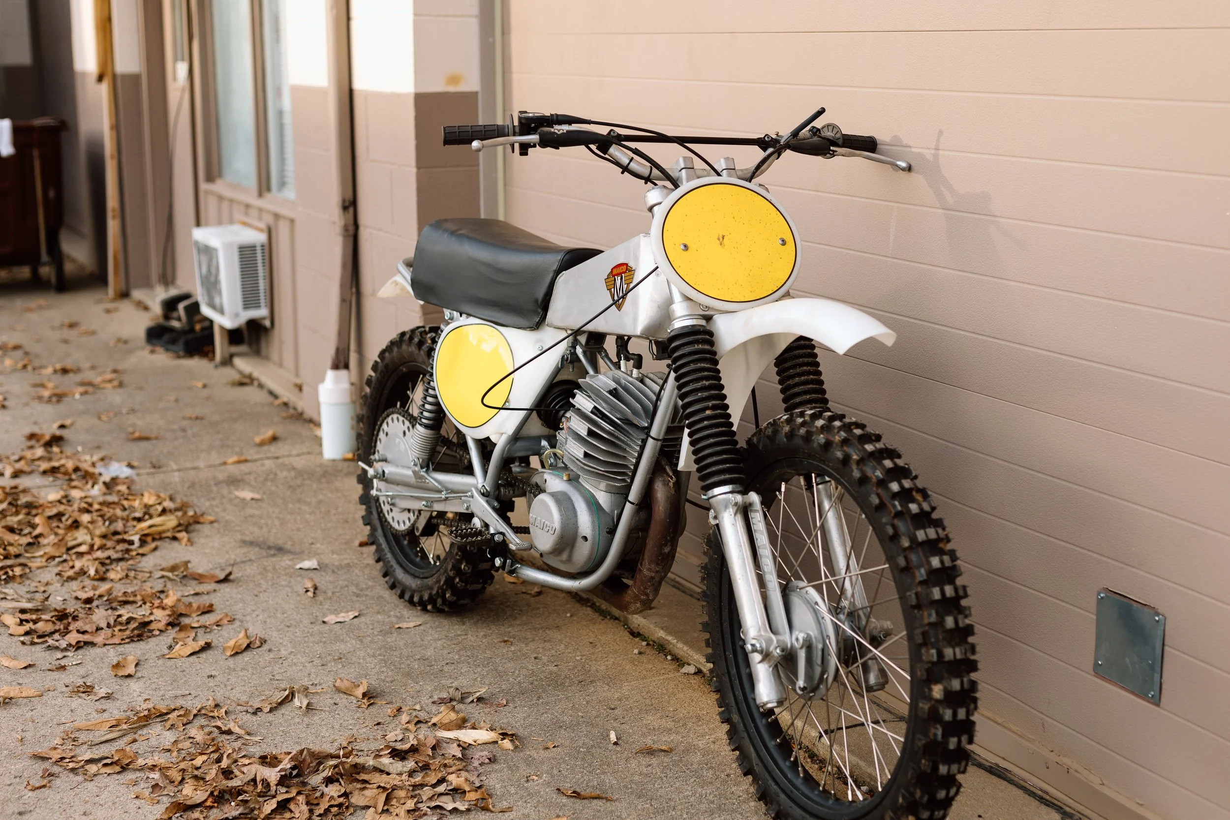 A vintage off-road motorcycle with yellow plates and a black seat, parked outdoors against a beige wall on a concrete sidewalk with fallen leaves around.