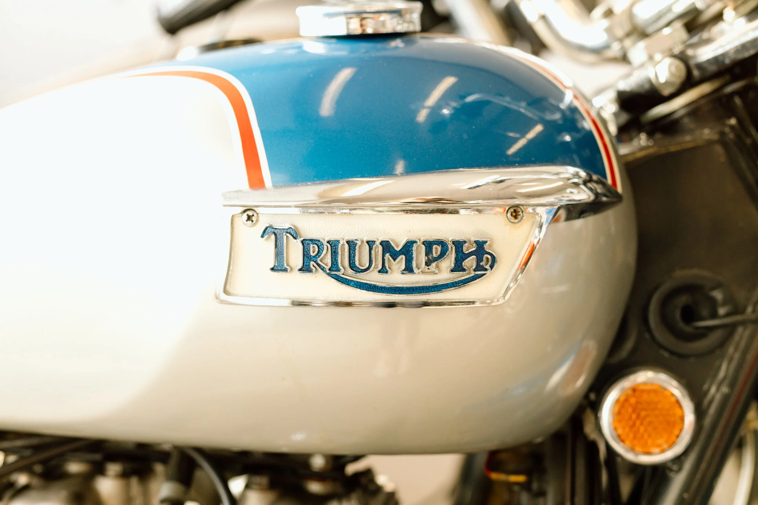 Close-up of a vintage Triumph motorcycle fuel tank with a blue, white, and orange paint scheme, and a Triumph emblem.
