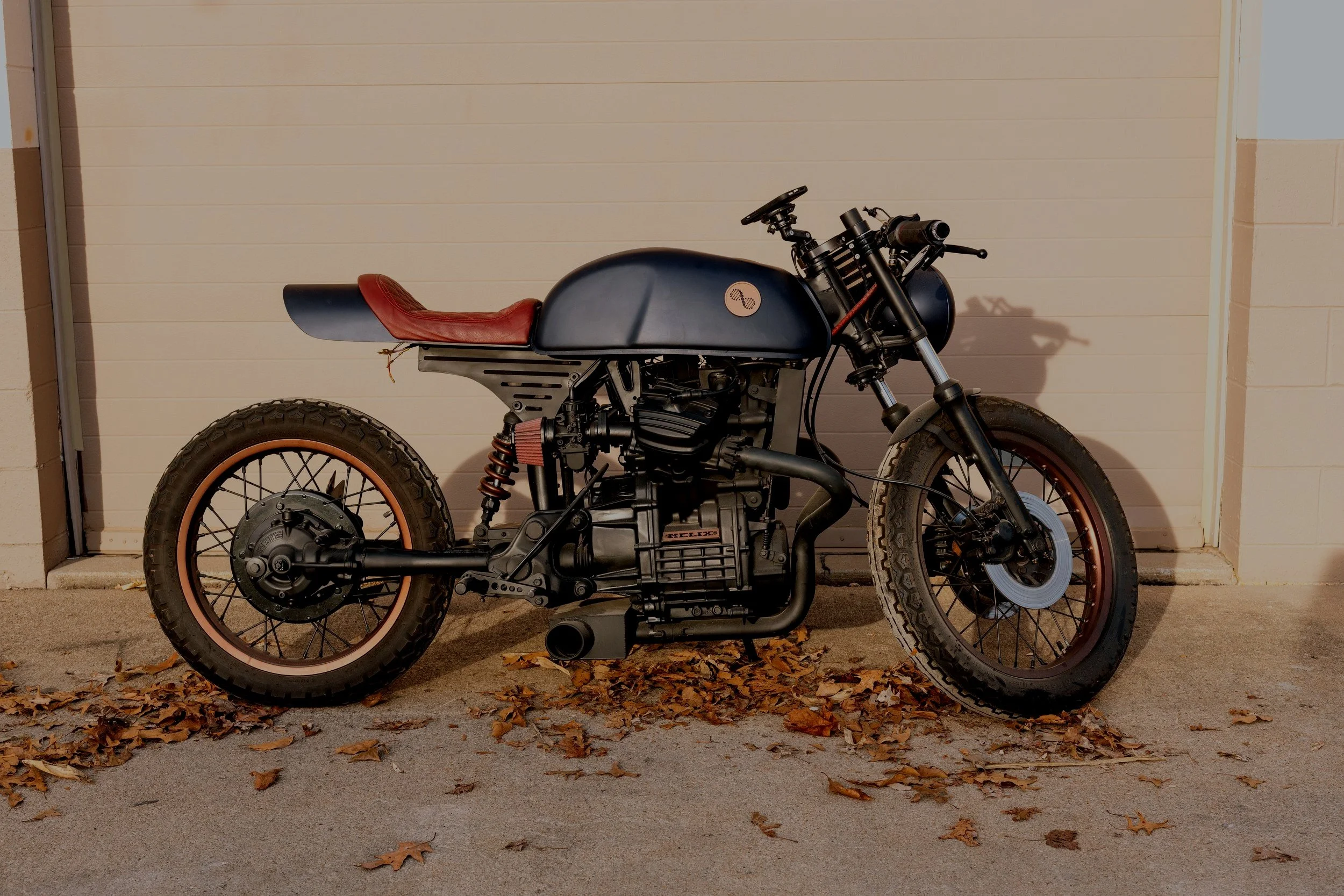 A custom-built motorcycle with a black and red color scheme parked on a concrete driveway with fallen autumn leaves, in front of a white garage door.