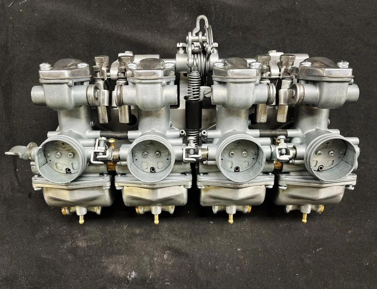 A vintage motorcycle carburetor assembly with four carburetors connected by linkages and springs, set against a black background.