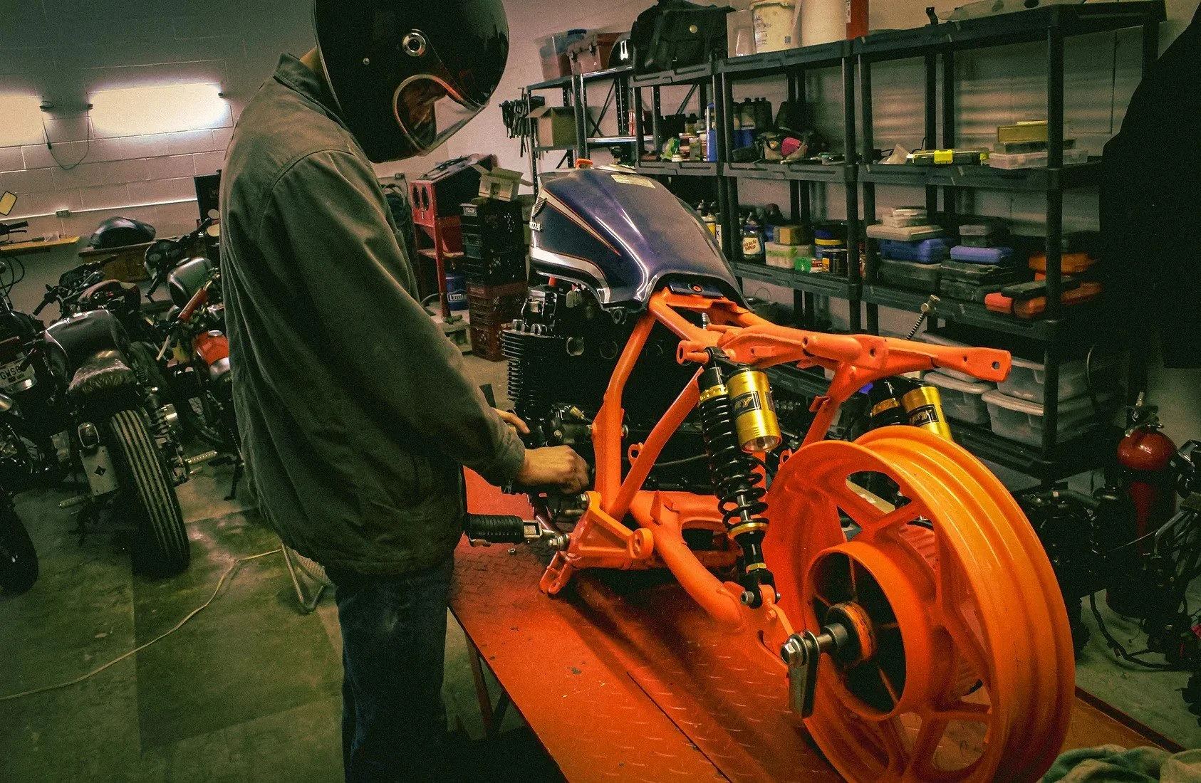A person wearing a helmet and gray jacket working on an orange motorcycle frame in a garage or workshop surrounded by tools and motorcycle parts.