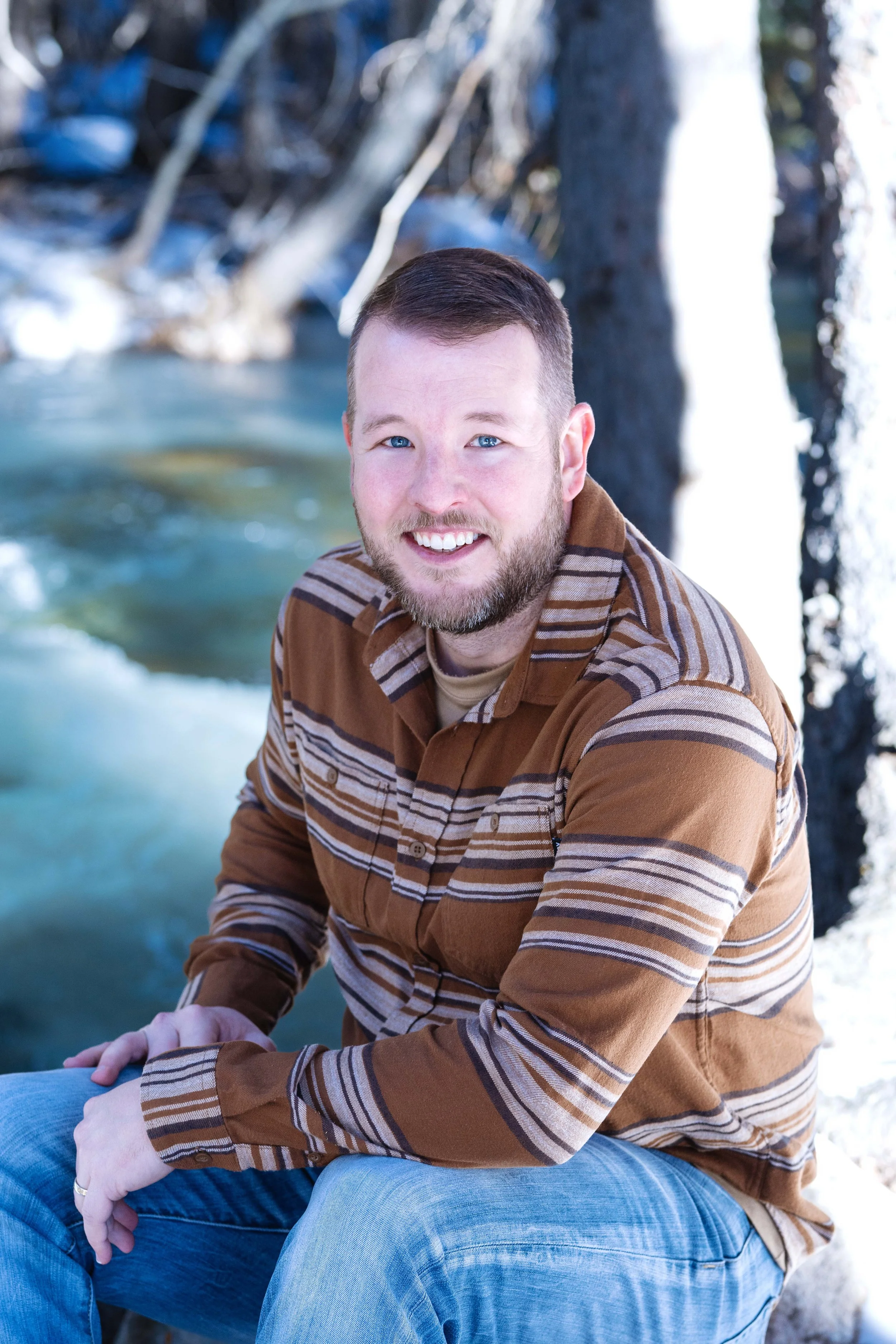 A man with short hair and a beard, smiling, wearing a brown striped shirt and light blue jeans, sitting outdoors near a flowing river with snow-covered trees in the background.
