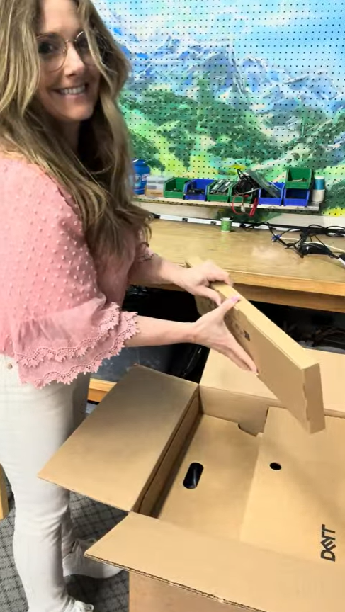 A woman with long, wavy blonde hair, wearing glasses, a pink blouse with lace sleeves, and white pants, is opening a cardboard box in a workshop or classroom setting, with tools and equipment on a table behind her and a colorful mountain landscape mural on the wall.