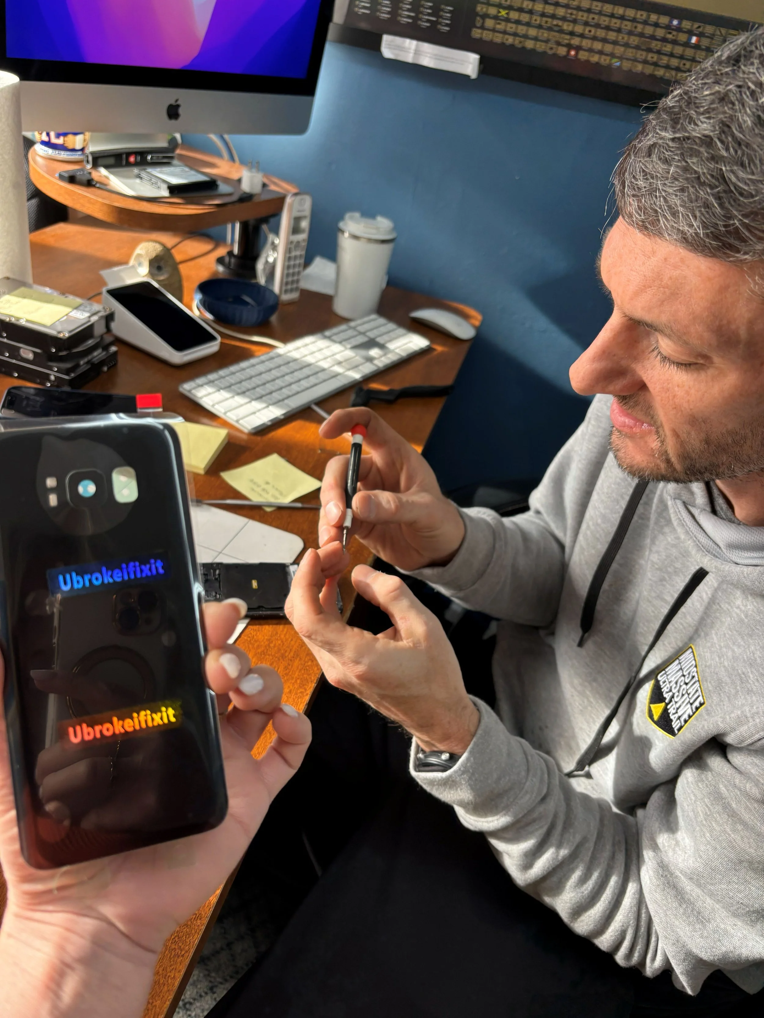A man is repairing a smartphone at a cluttered desk while another person holds a different smartphone showing the message 'Ubrokeifixit' in blue and red text.