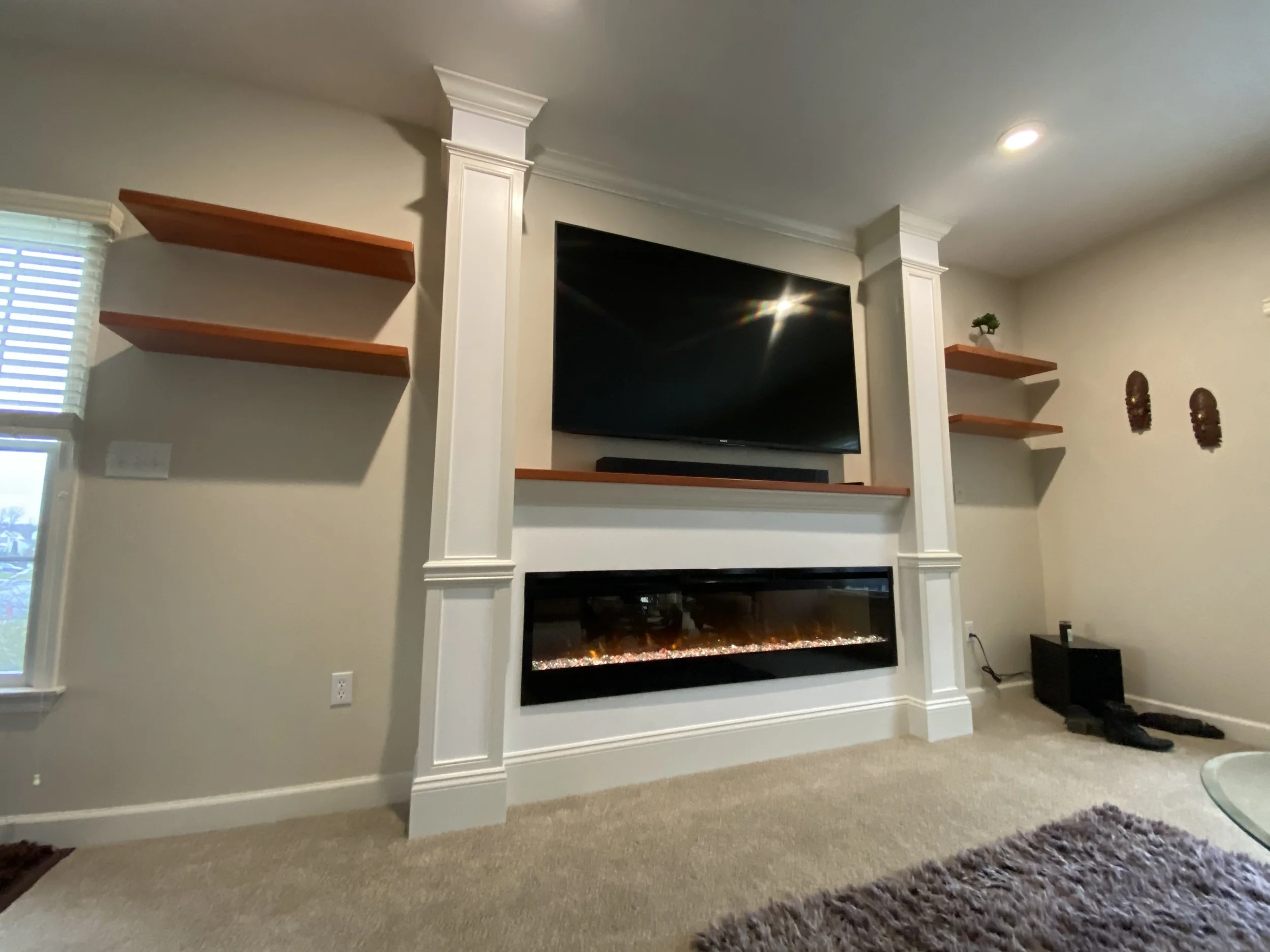 Living room with a wall-mounted flat-screen TV above an electric fireplace with a black frame, surrounded by white-built columns and beige walls. There are wooden shelves on either side of the TV, some decorated with small plants and masks. A window 