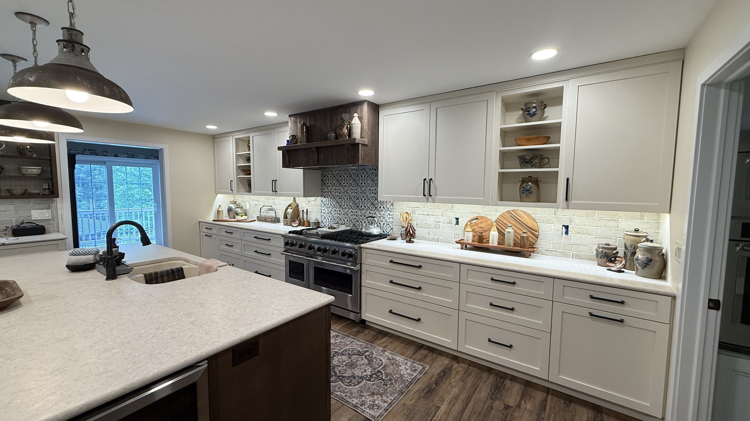 Modern kitchen with white cabinets, black handles, and a beige countertop. Features a stovetop with oven, decorative plates, and jars on the counter. Open shelving displaying dishes and decorative items. Pots and pans on the stove, sliding glass door