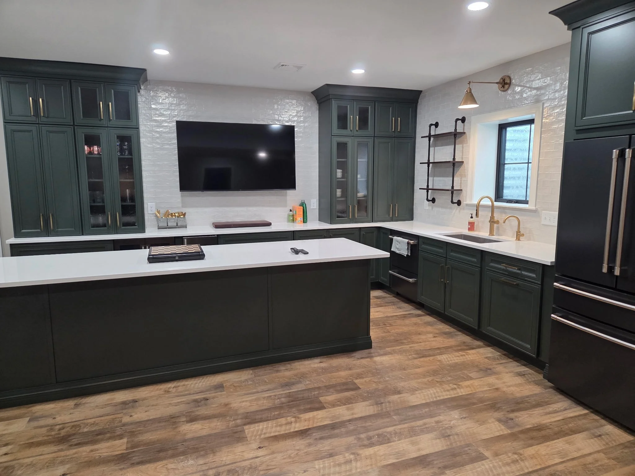 Modern kitchen with dark green cabinets, white countertops, a large black refrigerator, and a wood floor. There's a wall-mounted TV above the counter, and a window with wall-mounted light fixtures.
