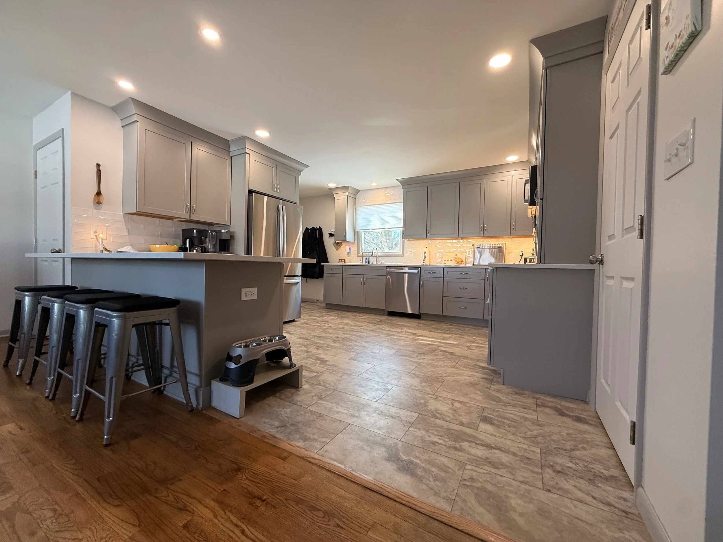 A modern kitchen with gray cabinets, stainless steel appliances, and a granite countertop. There are recessed lighting fixtures on the ceiling, a window above the sink, and a breakfast bar with three metal stools. The floor is a mix of wood and tile.