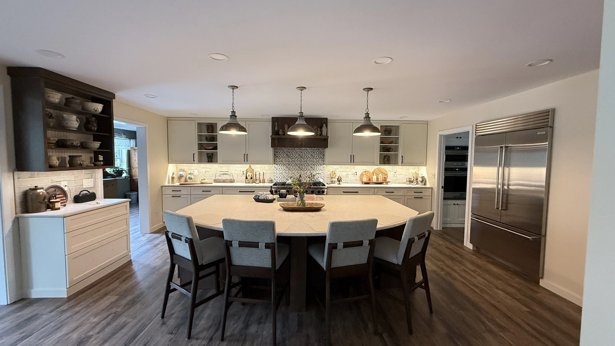 Modern kitchen with a large central island, beige chairs, white and gray cabinets, pendant lights, stainless steel refrigerator, and decorative items.