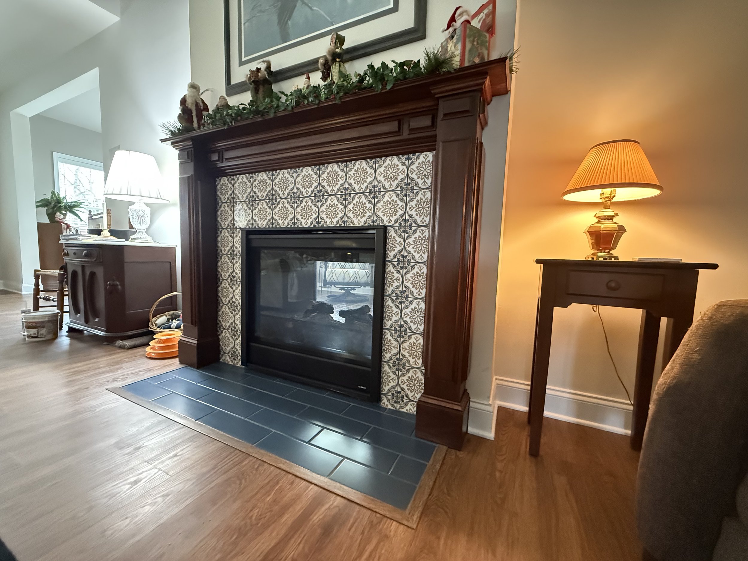 Living room with a decorative fireplace, Christmas garland, and four stuffed reindeer on the mantelpiece. To the right, a side table with a lit yellow lamp, near a beige armchair. To the left, a cabinet with a lamp and potted plant, and some stacked 
