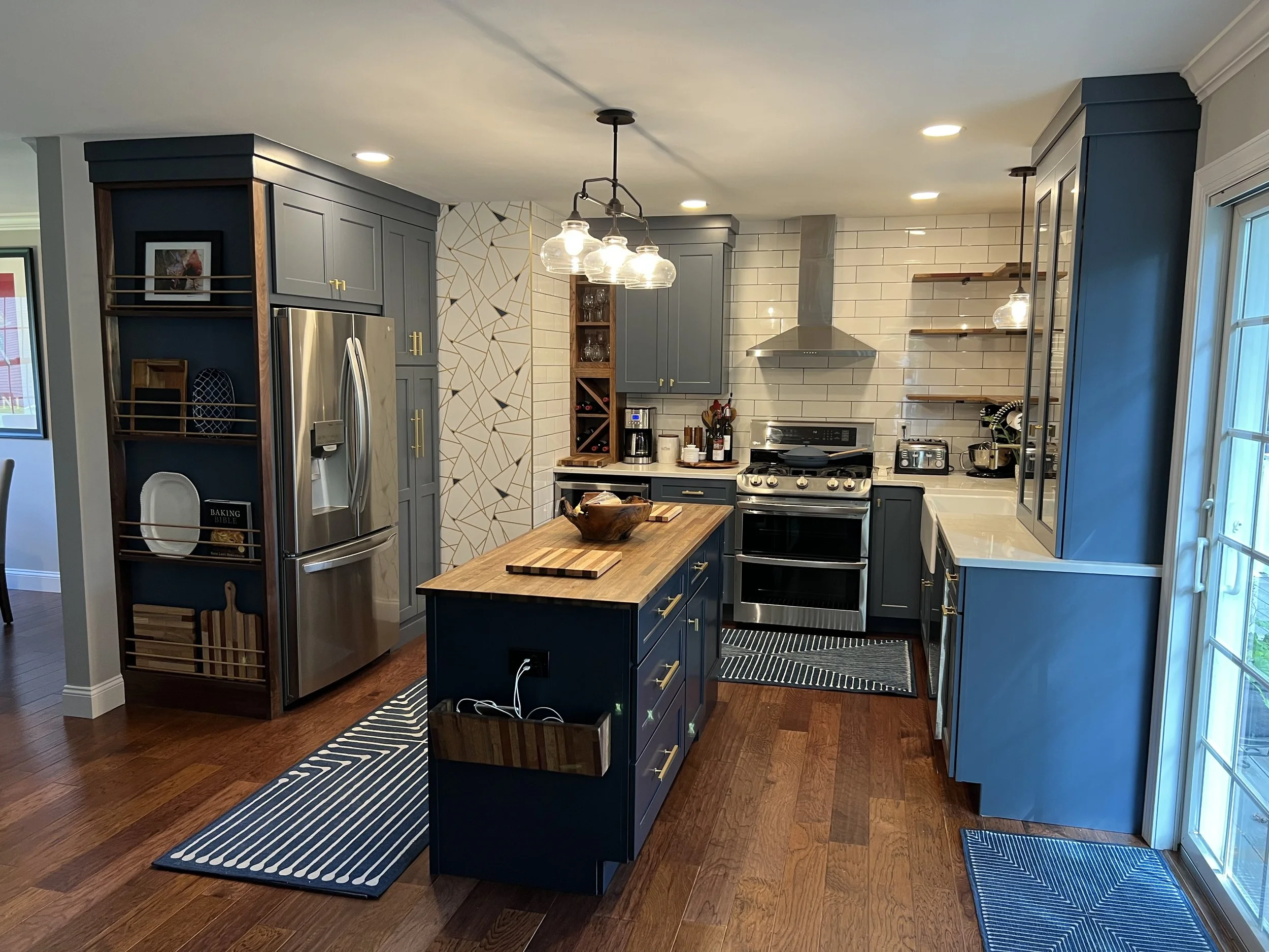 Modern kitchen with dark blue cabinets, wooden countertops, stainless steel appliances, open shelving, and a kitchen island with a wooden bowl. There are pendant lights overhead and a sliding glass door on the right.