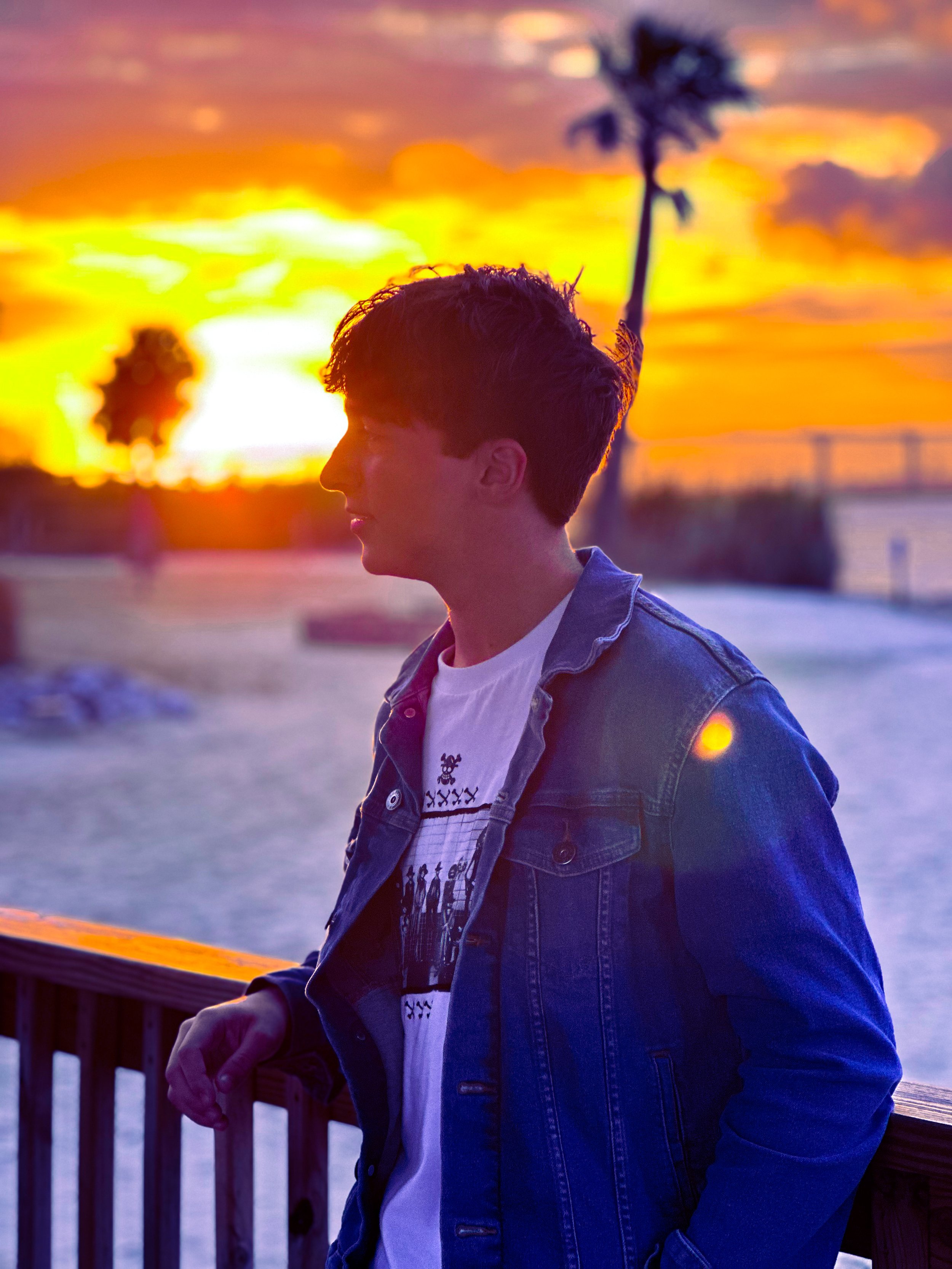 A young man with short hair wearing a denim jacket and a white graphic t-shirt, standing on a wooden deck during sunset with vibrant orange, yellow, and pink sky, and palm trees in the background.