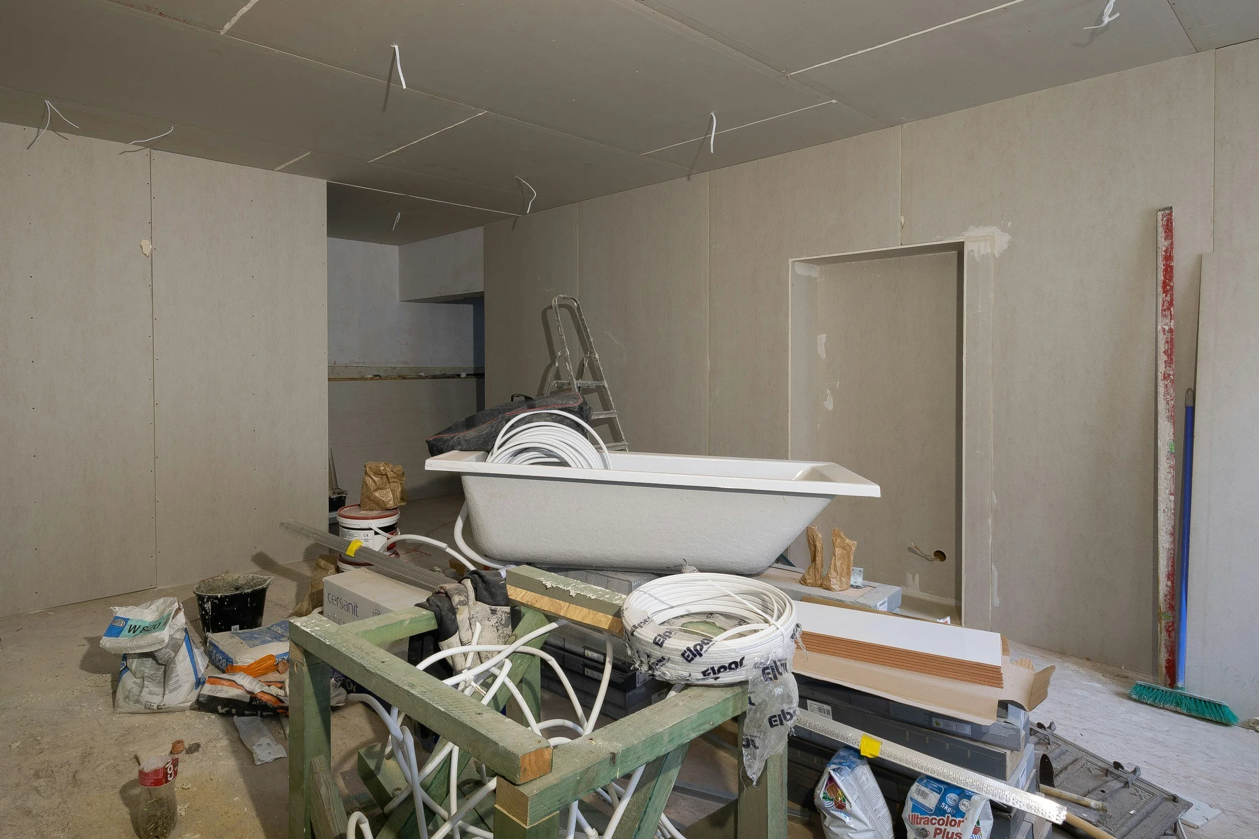 Construction site with unfinished walls, ceiling, and flooring; tools and building materials including a bathtub, ladder, pipes, and insulation.