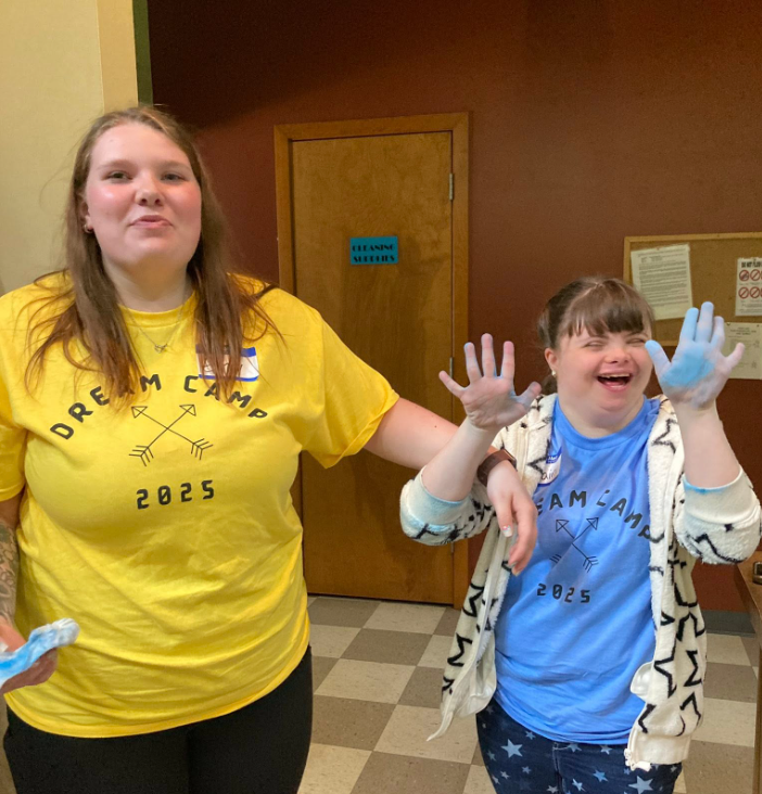 Two young girls smiling and playing in an indoor setting, wearing "Dream Camp 2025" t-shirts. One girl is in a yellow shirt, the other in a blue shirt with a hoodie, and they are interacting cheerfully.