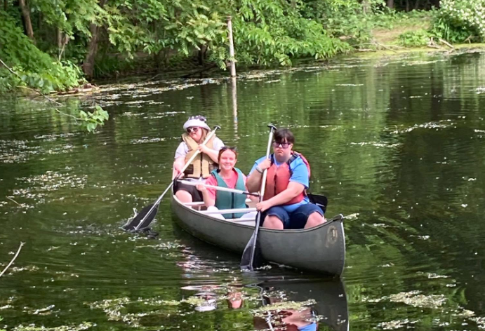 Three children in a canoe paddling on a calm river surrounded by green trees.