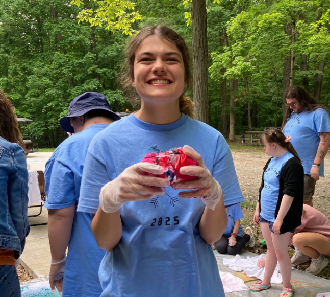 A young woman with brown hair smiling and holding a red and black tie-dye shirt at an outdoor gathering in a wooded area. She is wearing a light blue shirt and gloves, with several other people visible in the background, some wearing similar shirts.