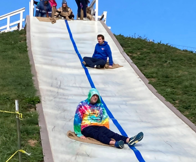 A person in a tie-dye hoodie sliding down a toboggan slide with a blue line painted on it, while another person in a blue jacket sits on the slide, and several people at the top of the slide prepare to go down.