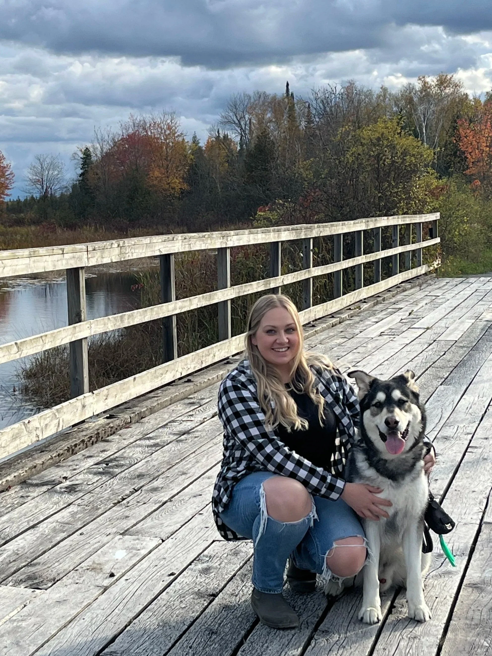 A woman with long blonde hair, wearing a plaid shirt and ripped jeans, kneeling on a wooden bridge, holding a smiling Siberian Husky dog, with autumn trees and a cloudy sky in the background.