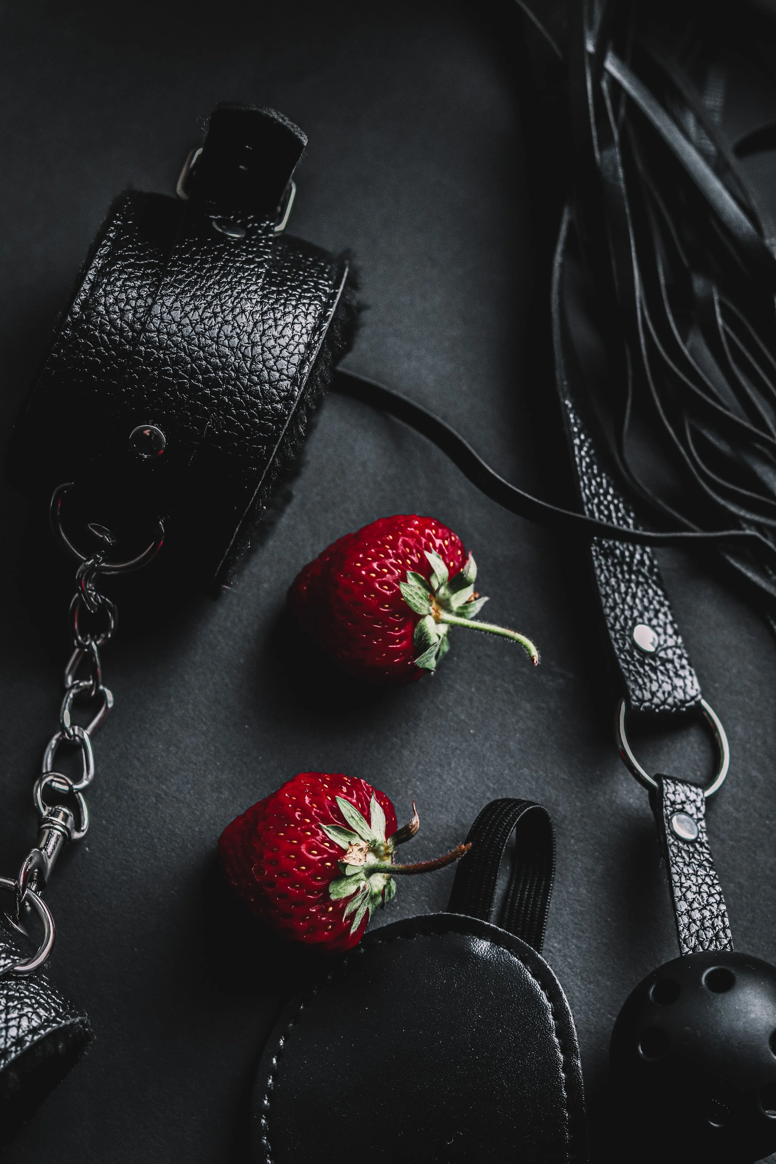 Close-up of a black textured leather camera strap with a silver chain linking the strap to a camera, two strawberries with green leaves, and black photography gear on a black surface.