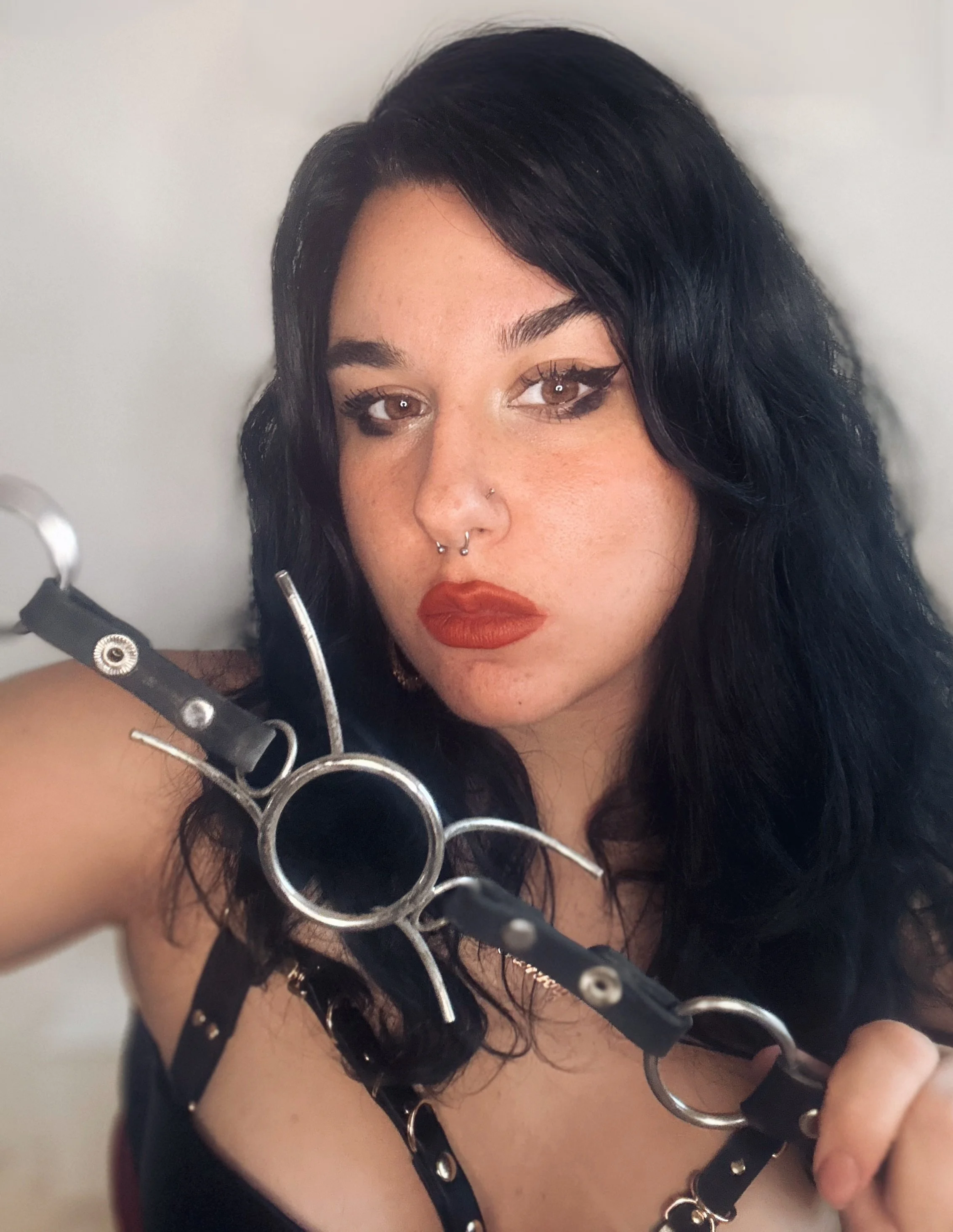 A woman with dark hair, wearing makeup, with a nose piercing, dressed in black leather with metal rings, holding a collection of metallic curled objects, taking a selfie against a plain light-colored background.