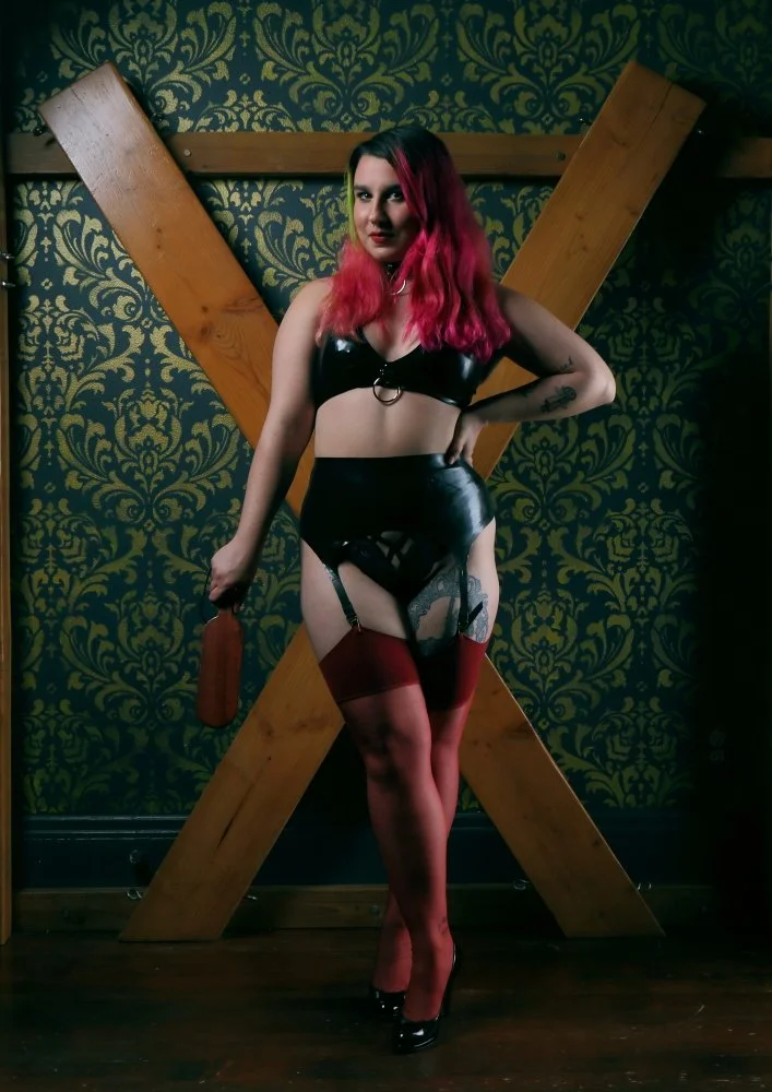 Woman with pink hair in black leather lingerie and red stockings standing in front of a large wooden cross mounted on a wall with ornate green wallpaper.