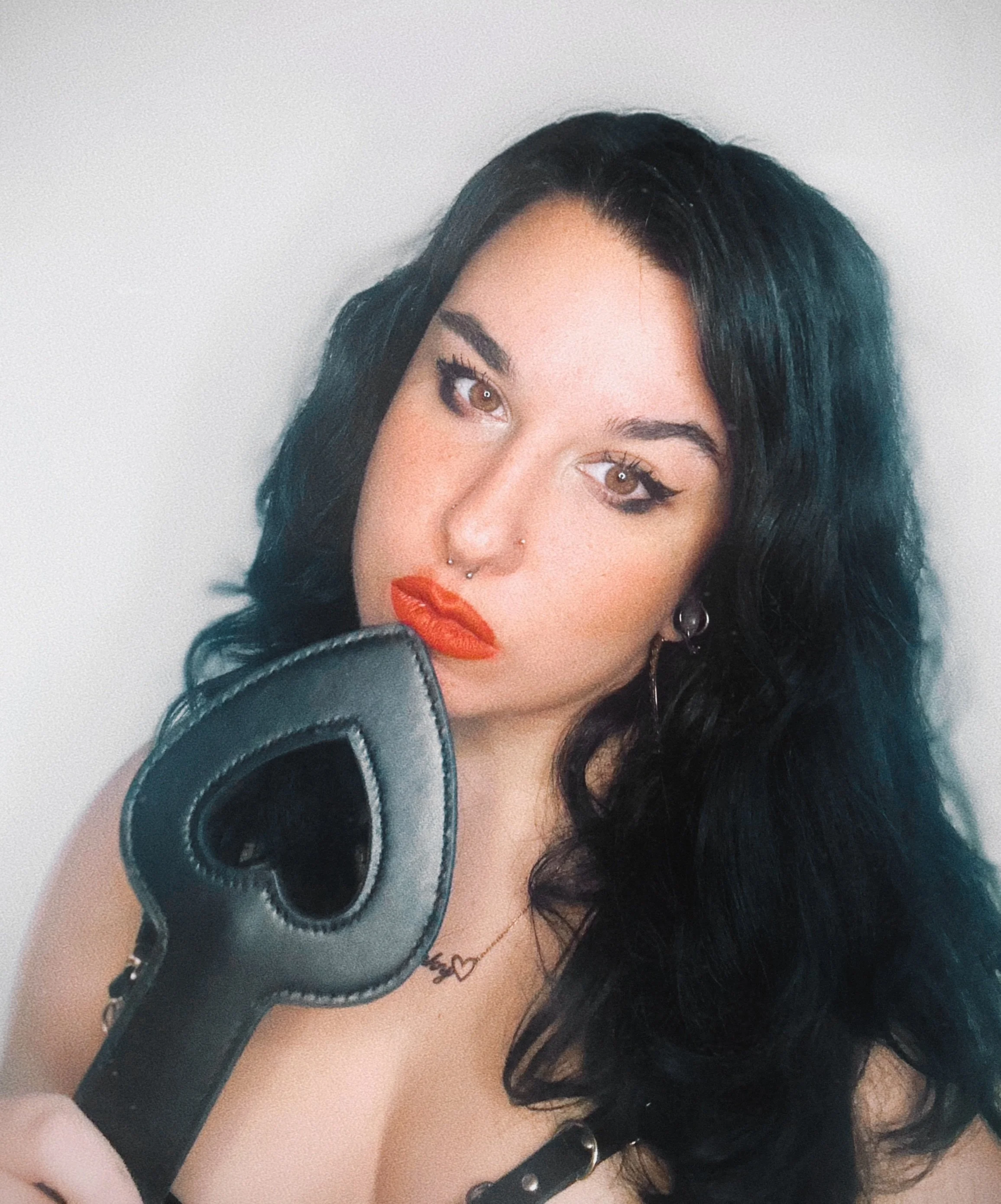 A woman with dark hair, makeup, and piercings holding a black leather object close to her face, partially covering her chin and shoulder, with a plain light background.