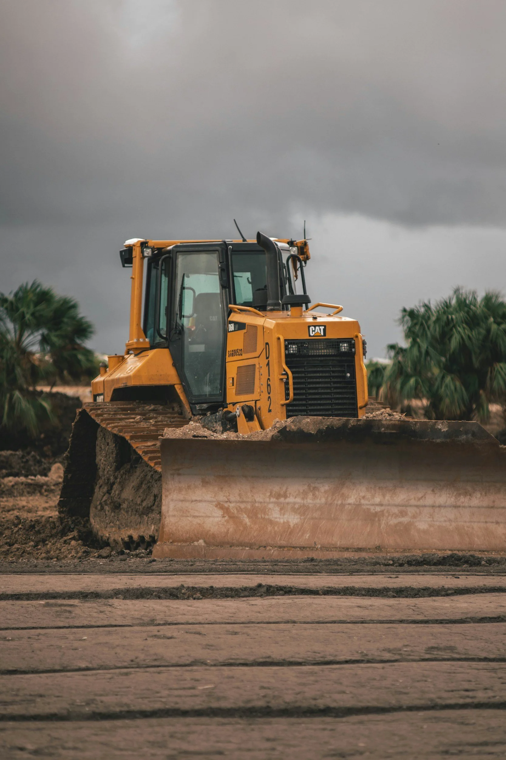 A yellow bulldozer working on a construction site with dark storm clouds overhead and palm trees in the background.