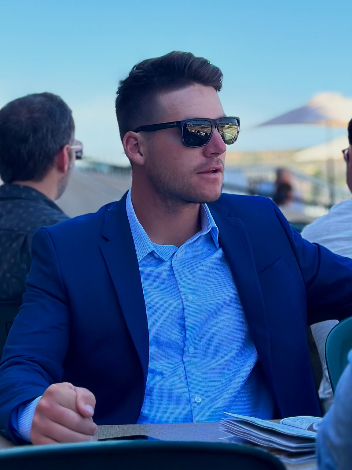 A man wearing sunglasses, a blue blazer, and a light blue dress shirt sits at an outdoor event with umbrellas and other people in the background.