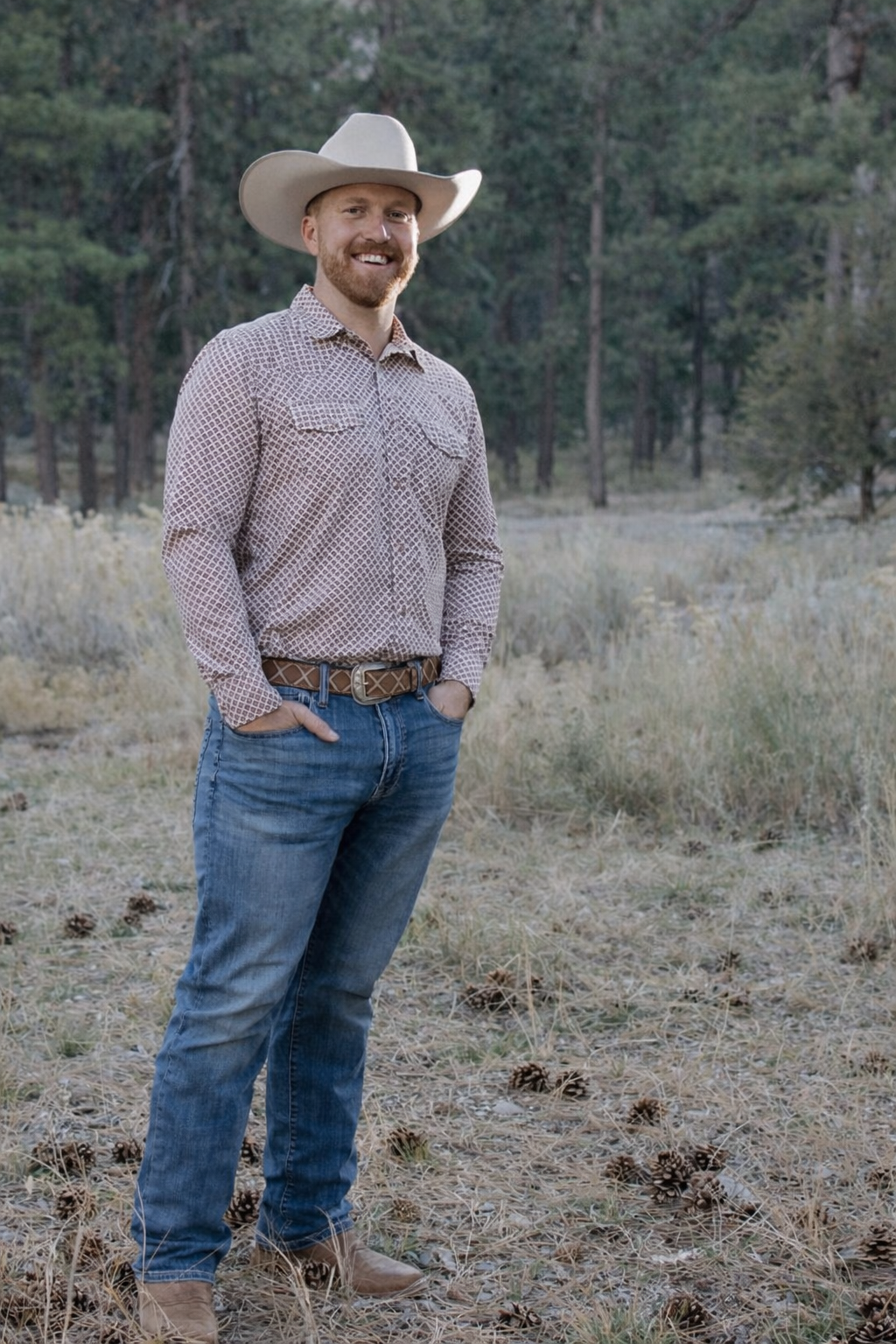 A man in a cowboy hat and patterned shirt standing in a forest clearing with pine trees in the background.