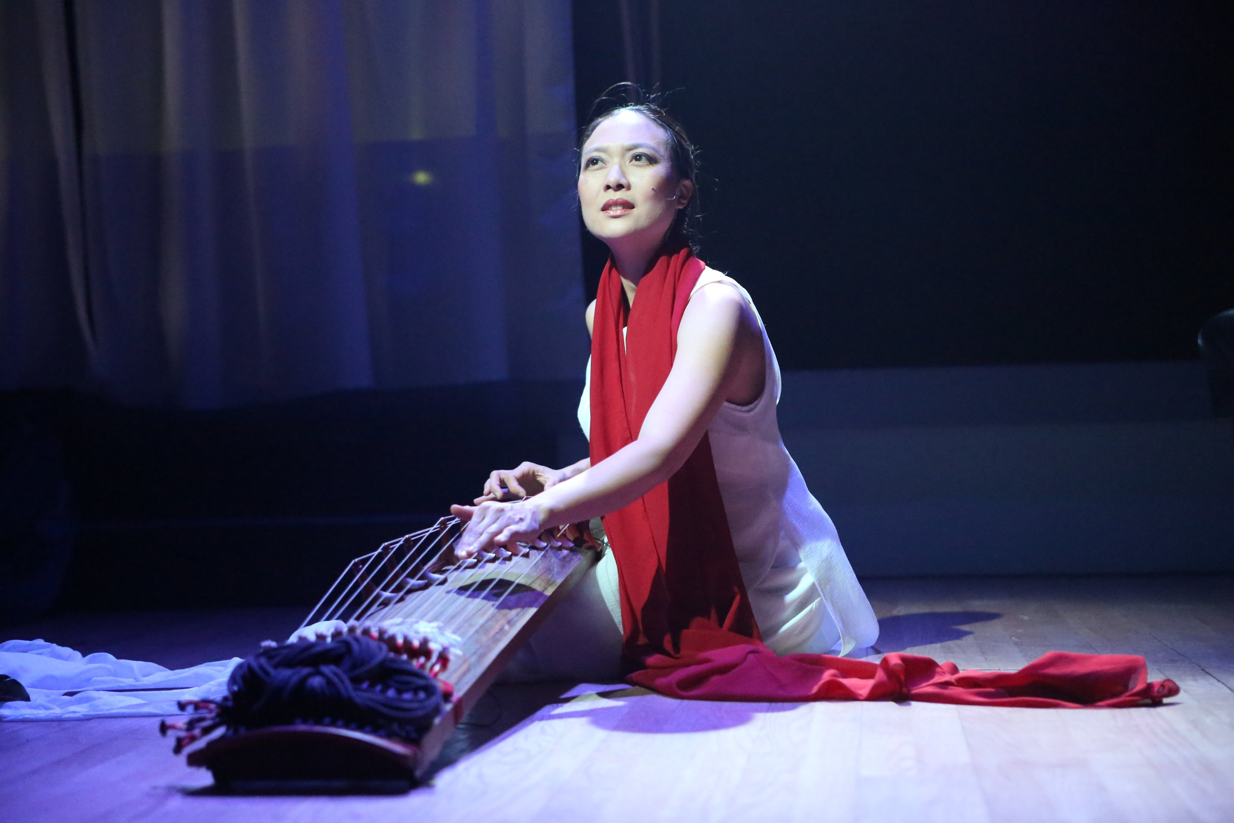 A woman dressed in white kneels on a stage, playing a zither-like musical instrument with a red scarf draped around her neck, under stage lighting.