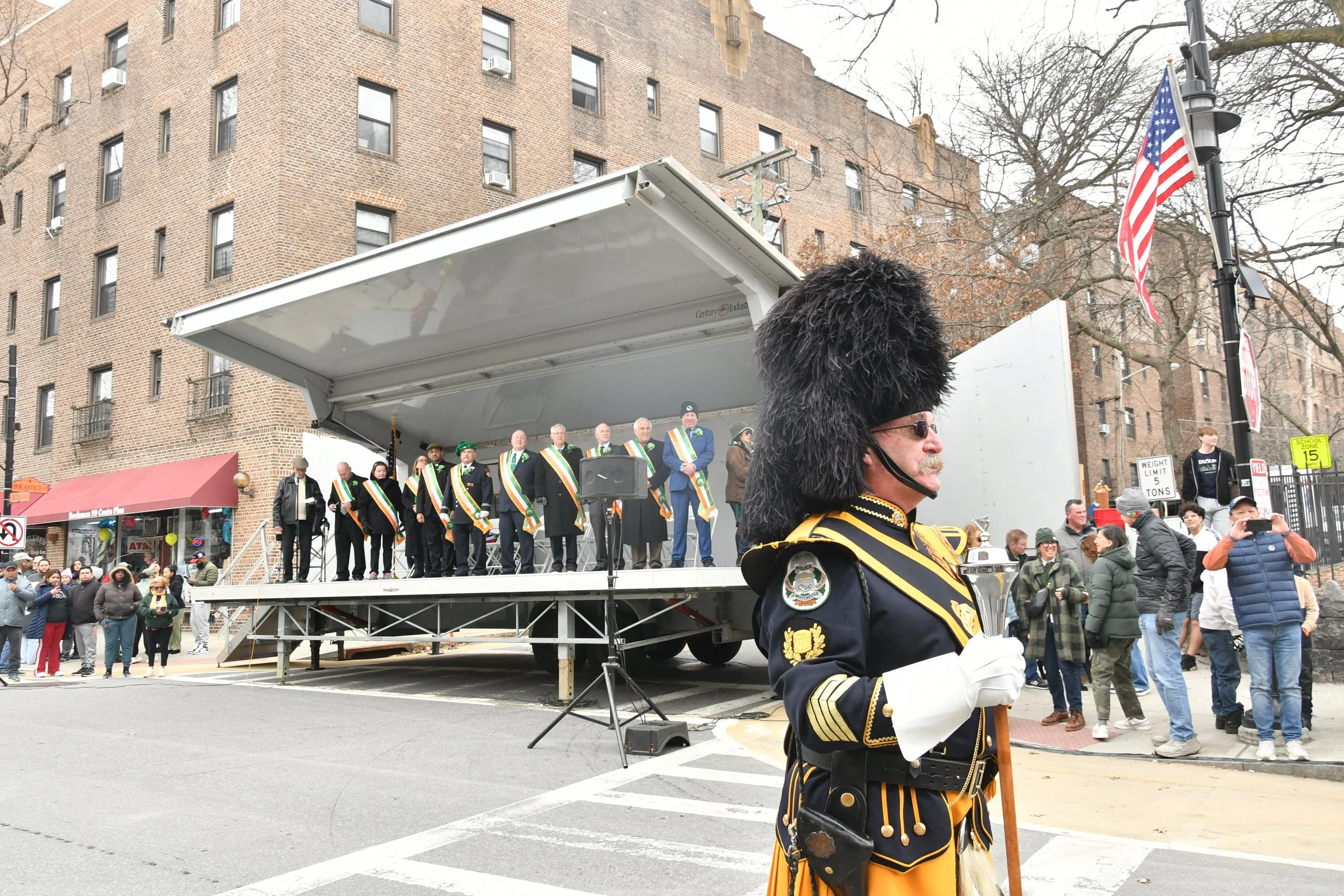 A parade scene showing a crowd gathered around a parade float stage with a group of men in formal attire and sashes, with a man in a historic military uniform standing in the foreground. The stage is on a city street surrounded by buildings, trees, a