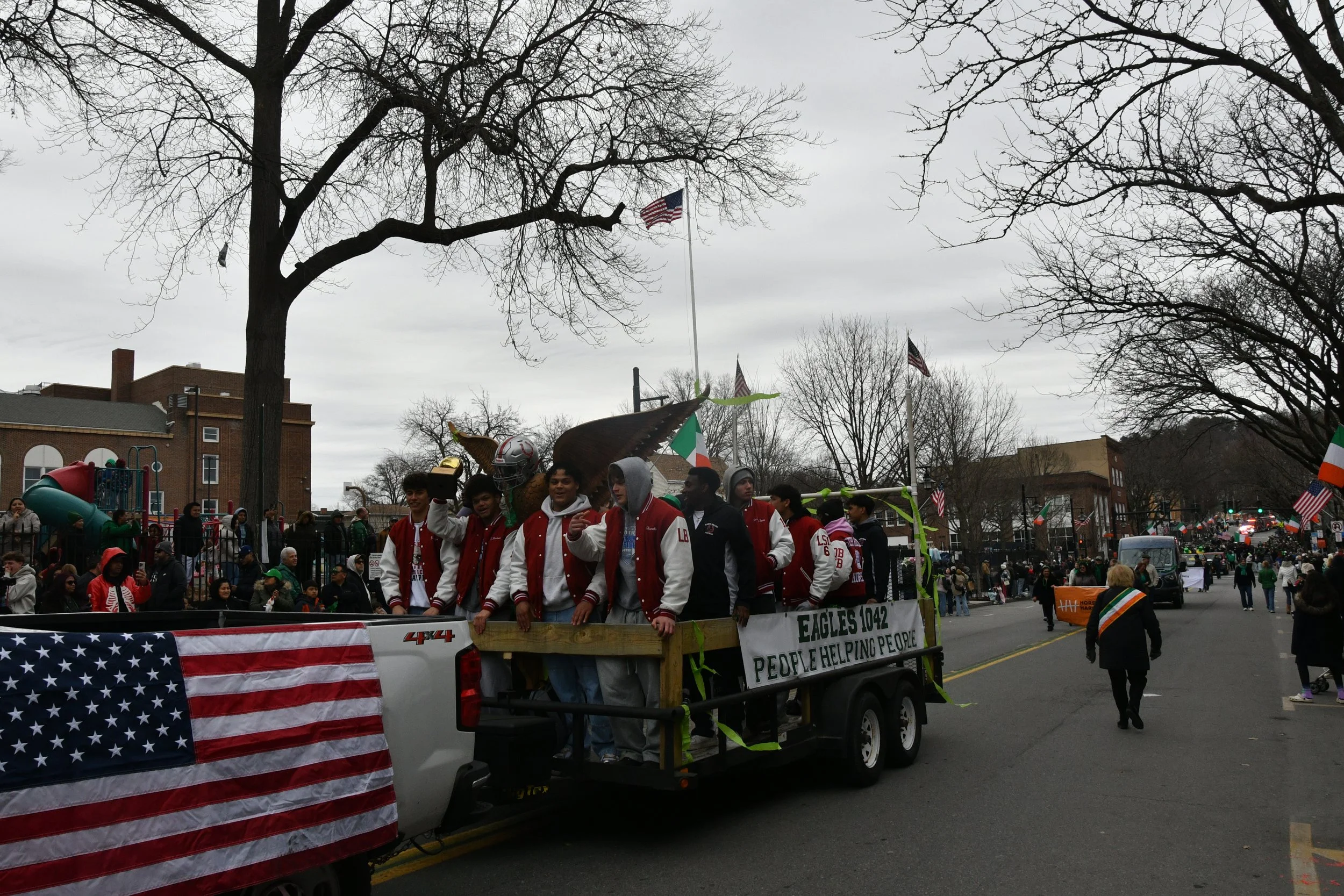 A parade float in a parade with a group of young people standing on it. The float has an American flag and a sign that reads 'EAGLES 1042 PEOPLE HELPING PEOPLE.' The background shows leafless trees, buildings, and more parade participants and spectat