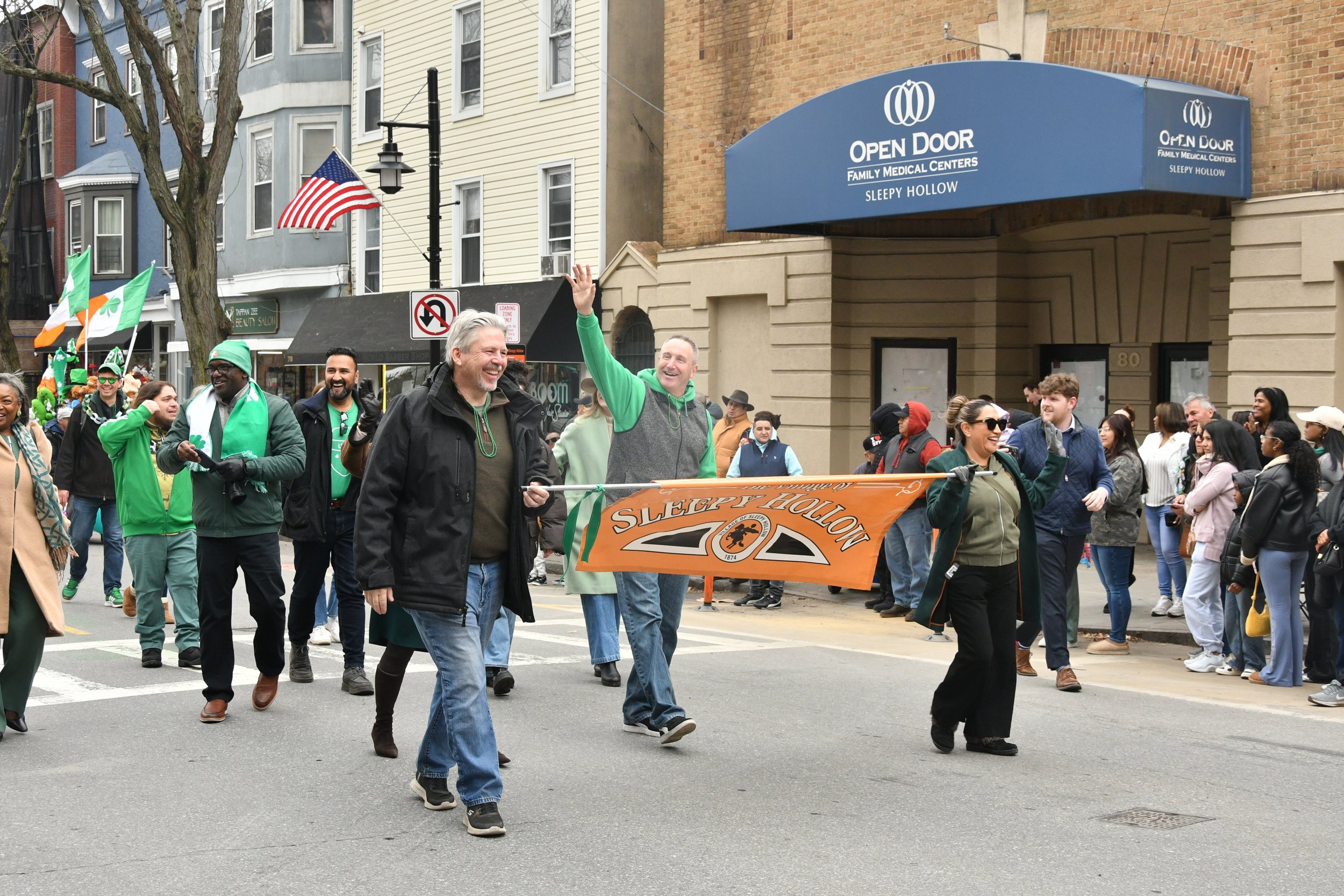 A parade or march with people smiling and waving, holding a banner for Sleepy Hollow, as they walk down a city street. Some participants wear green clothing and accessories, and flags with green, white, and orange colors are visible.