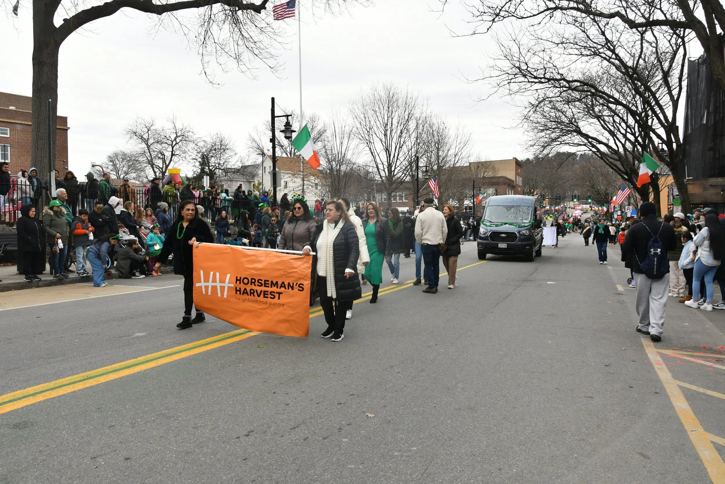 People participating in a parade or march on a city street, some holding a banner that reads 'Horseman's Harvest neighborhood pantry,' and American and Irish flags are visible, with onlookers lining the sidewalk.