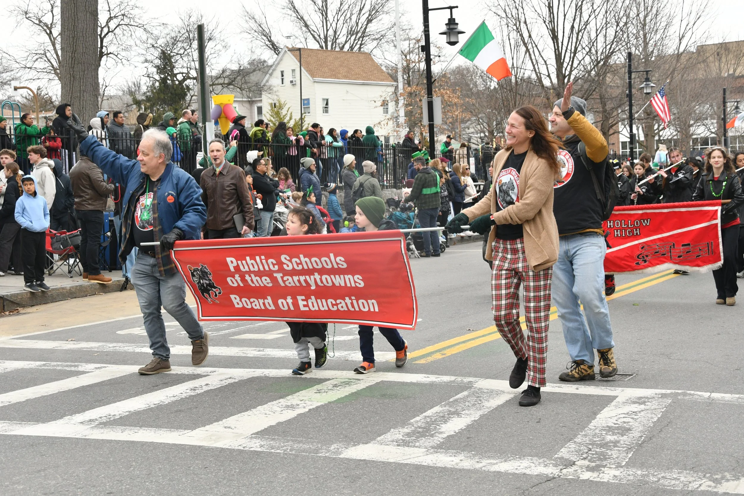 People participating in a parade or demonstration, holding a red banner with text about public schools, walking across a crosswalk on a city street. Spectators observe from the sidewalk and a fence, with leafless trees, houses, and streetlights in th