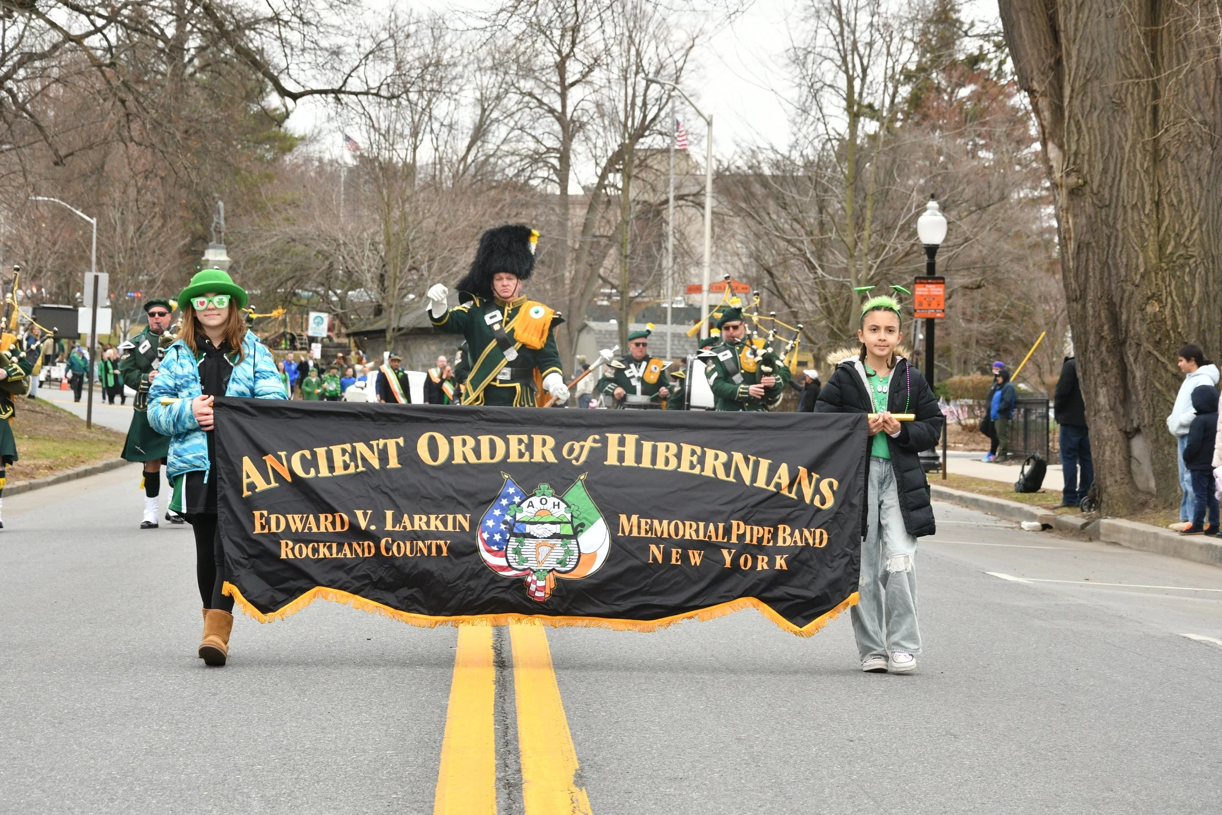 A parade with children holding a banner that reads 'Ancient Order of Hibernians, Edward V. Larkin Memorial Pipe Band, Rockland County, New York'. A man dressed in a black uniform with a tall black hat is leading the parade, along with children wearin