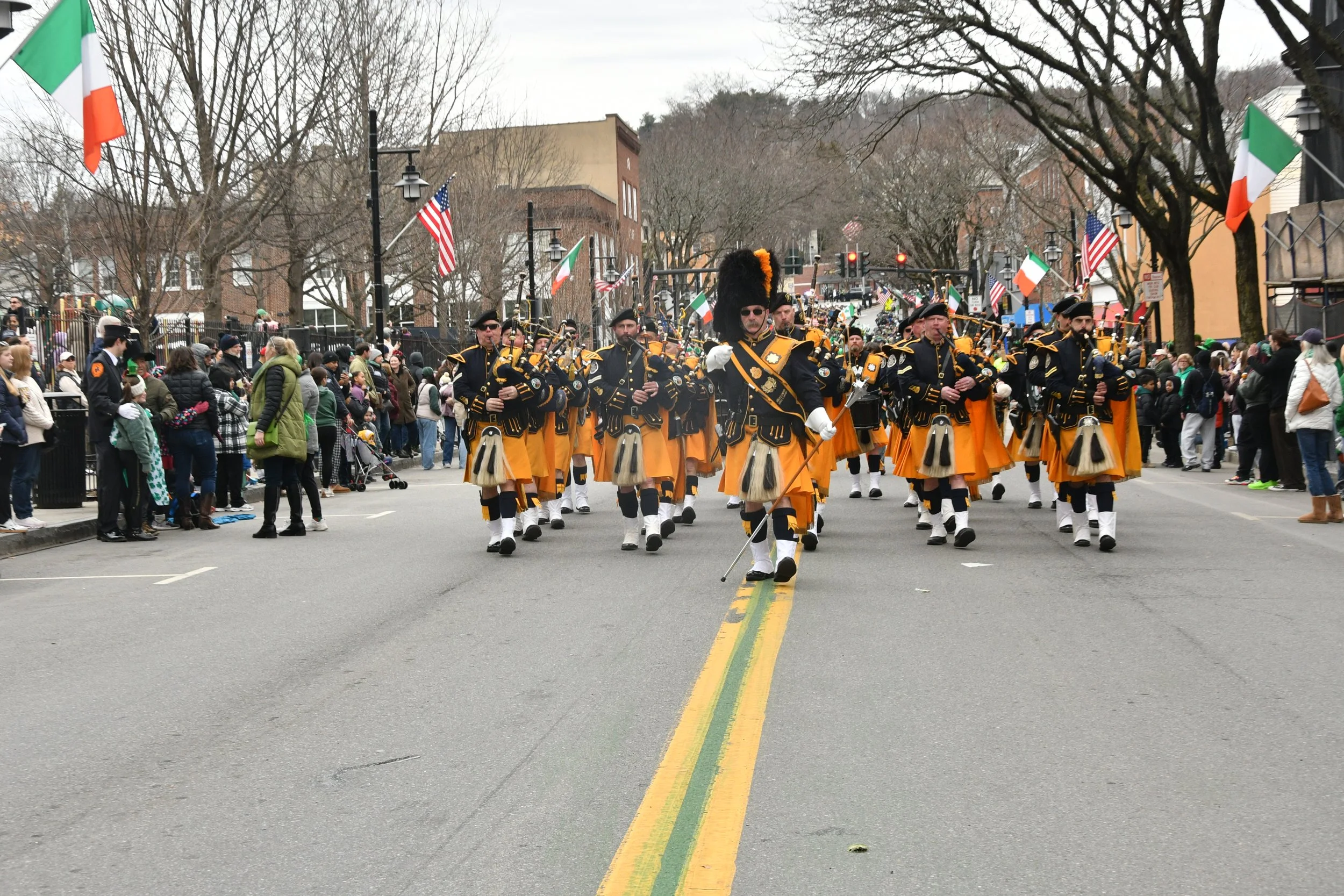 A parade with musicians dressed in yellow and black kilts, playing bagpipes and drums, marching down a city street lined with spectators and American and Irish flags, with leafless trees and buildings in the background.