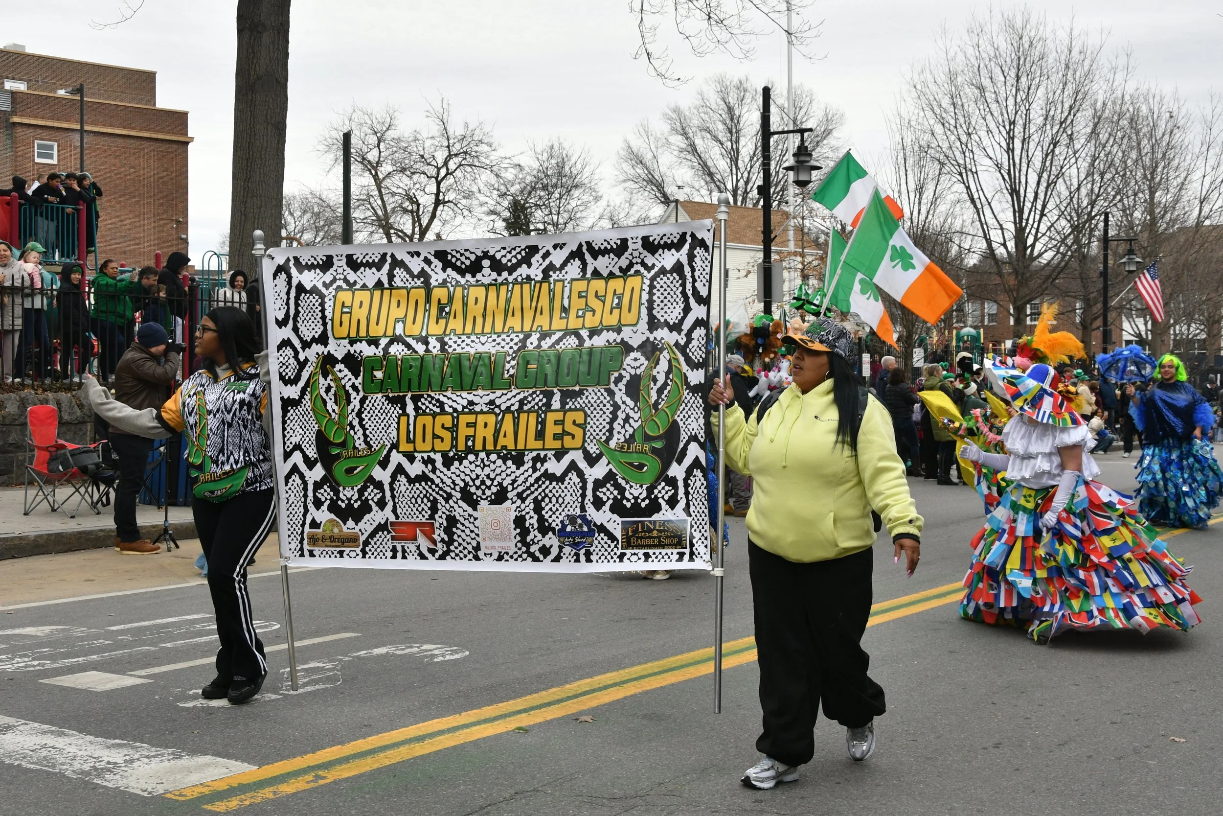 Two women are walking on a parade street, holding a banner that reads 'Grupo Carnaval Esc. Carnaval Group Los Frailes', with other parade participants dressed in colorful costumes and flags of Ireland and Ukraine, and spectators lining the sidewalk b