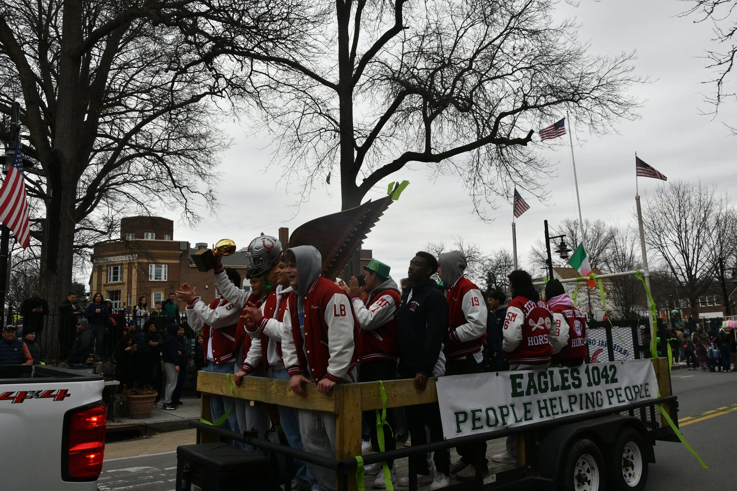 A group of people in red and white jackets participating in a parade, standing on a float with a sign that reads 'EAGLES 1042'. They are holding awards and smiling, with American flags and leafless trees in the background on a cloudy day.