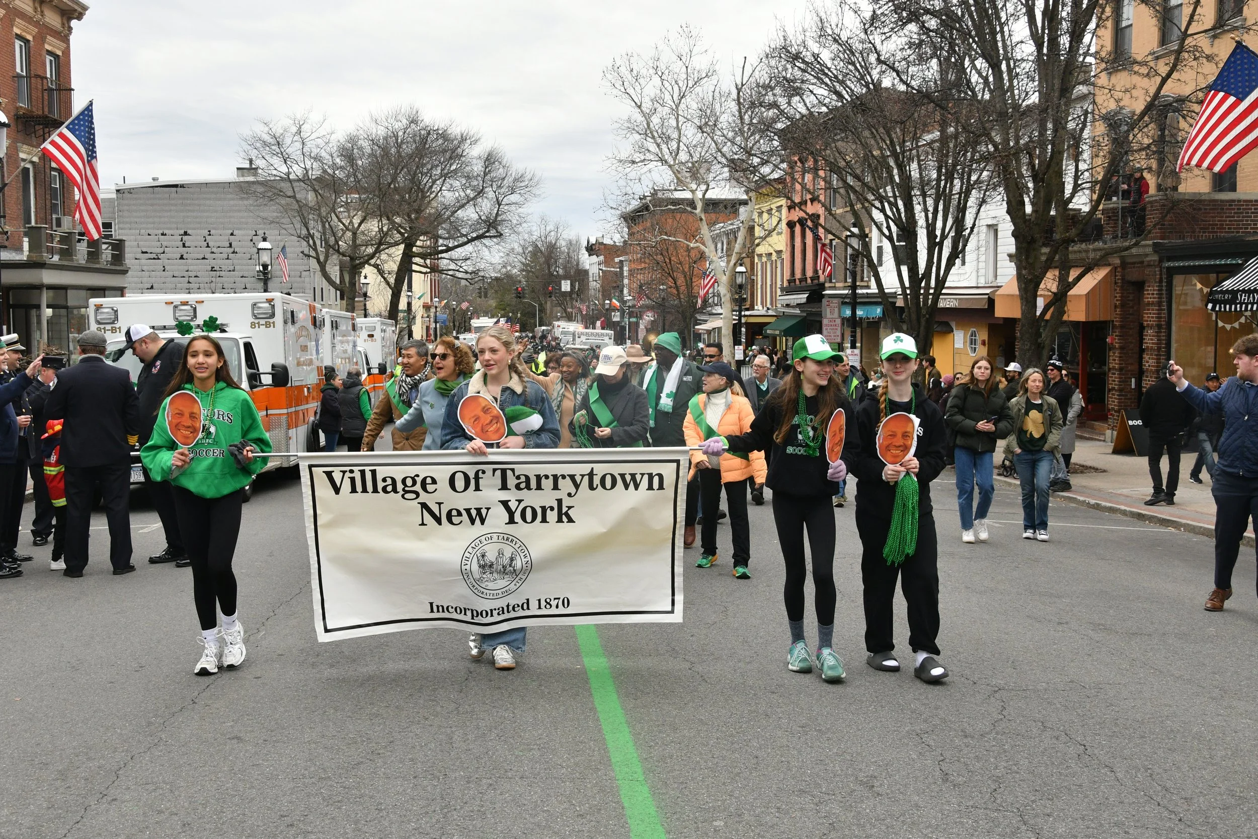 A parade with people walking down a street in Tarrytown, New York, carrying a banner that reads 'Village of Tarrytown, New York, Incorporated 1870.' The participants are dressed casually, some holding cardboard cutouts of a man's face, and are wearin
