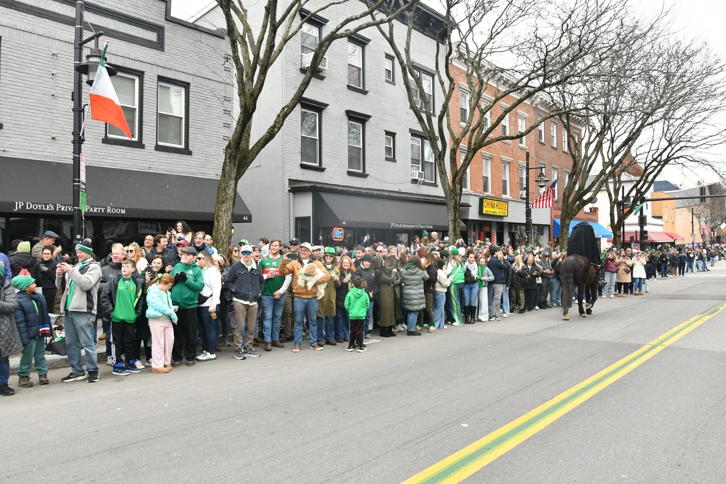 A large crowd of people gathered on a city sidewalk celebrating Saint Patrick's Day, some dressed in green, with some holding or wearing accessories like hats and scarves. There are buildings and leafless trees in the background, along with flags and
