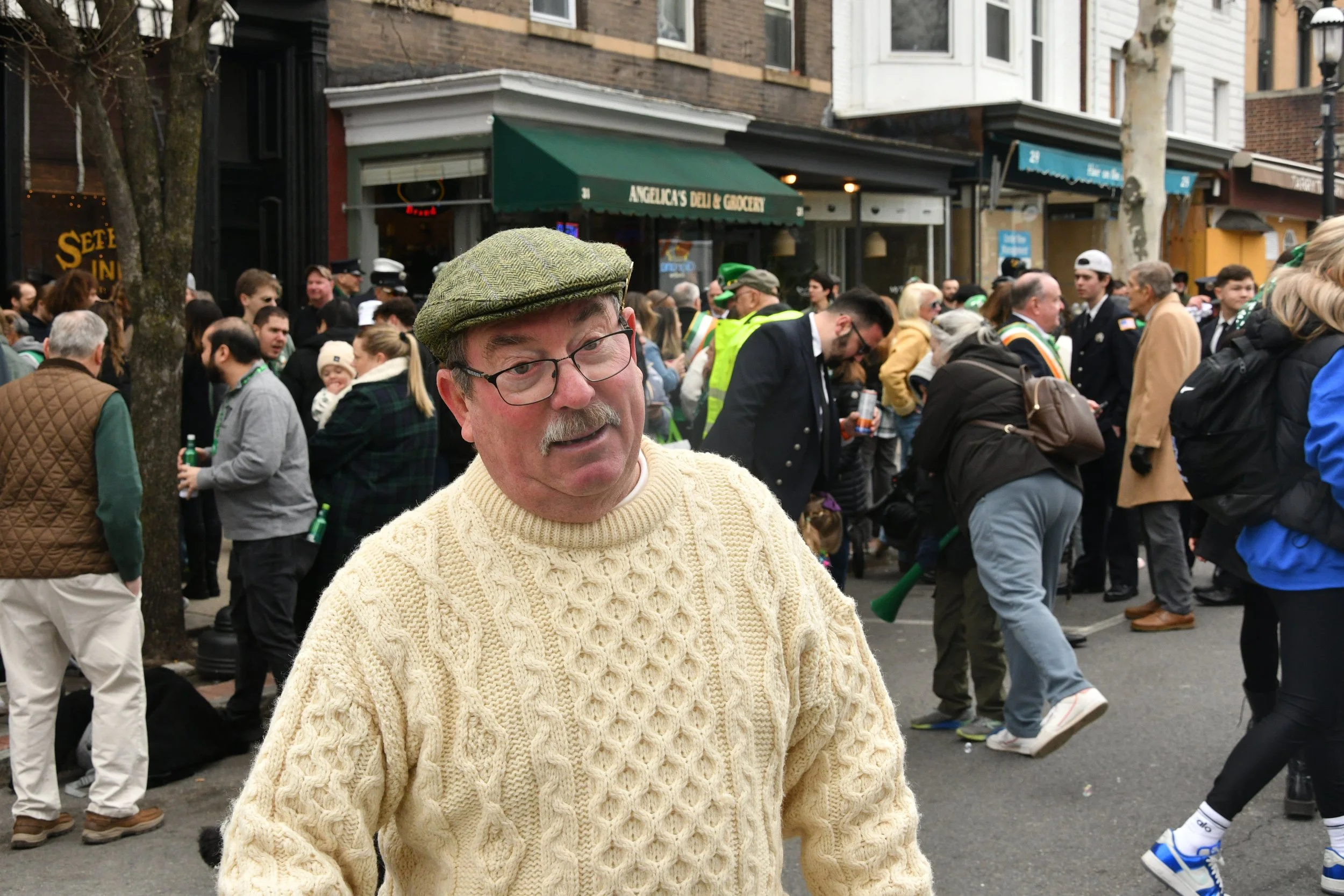 A man wearing glasses and a green flat cap standing in front of a crowd on a city street.
