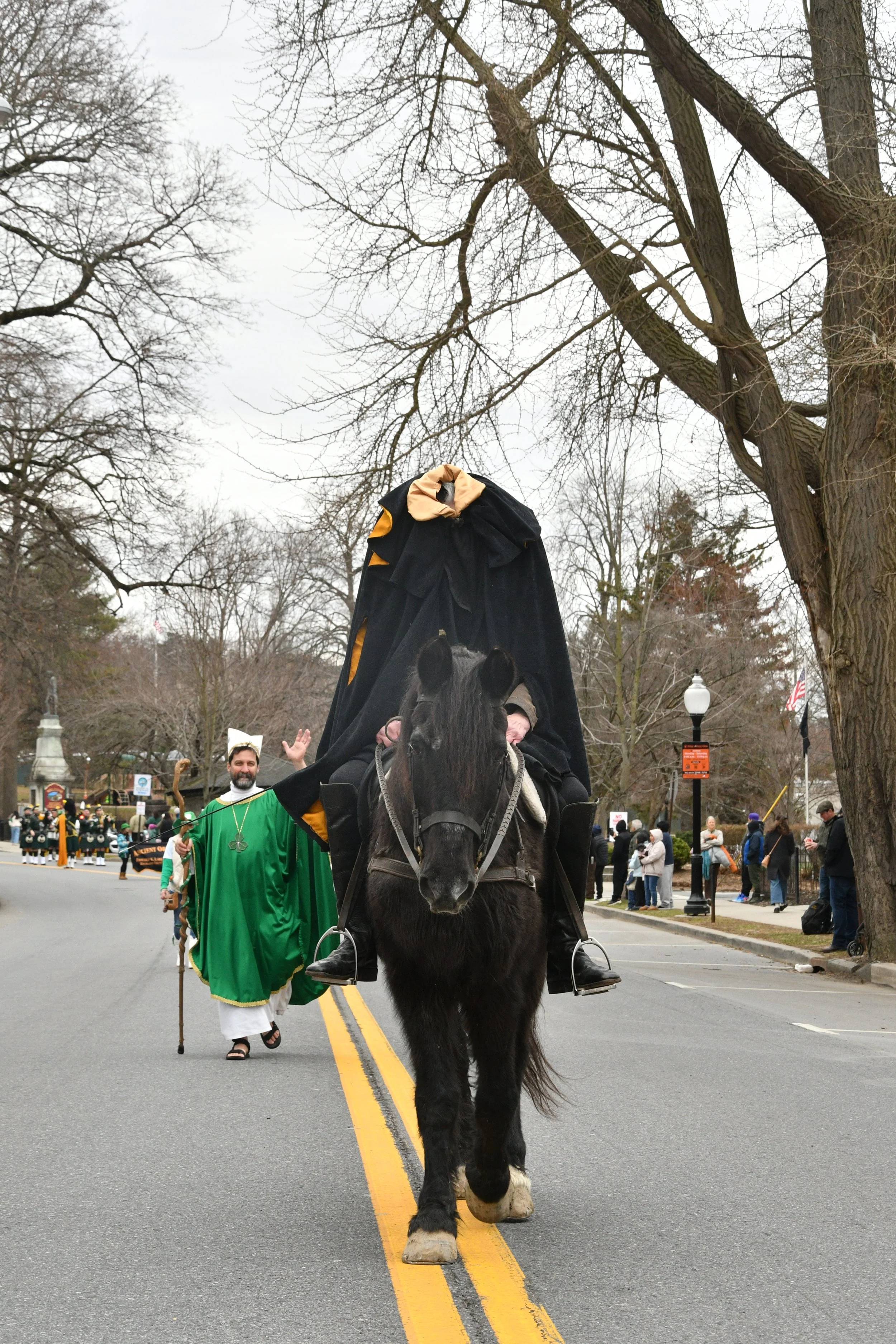 A person riding a black horse in a parade, holding a black flag, with a man in a green robe and mascot costume walking behind, and a crowd of spectators on the sidewalk and in the background.