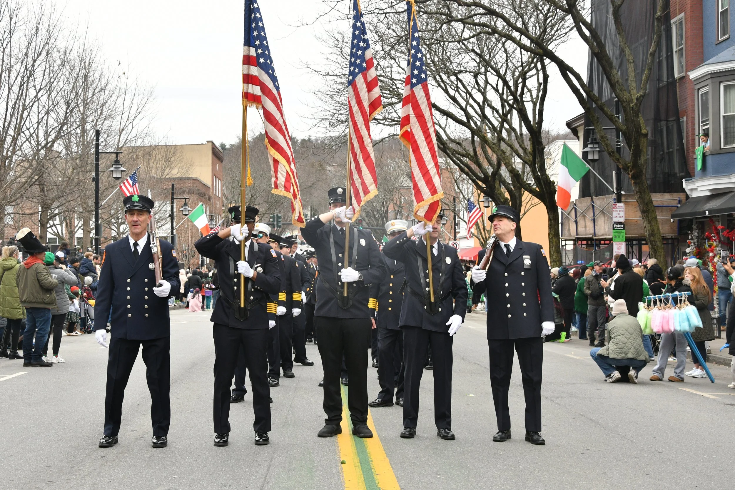 A parade float with uniformed individuals holding American flags, marching down a street. Onlookers watch from the sides of the street, with some participating in the celebration amid leafless trees and buildings.